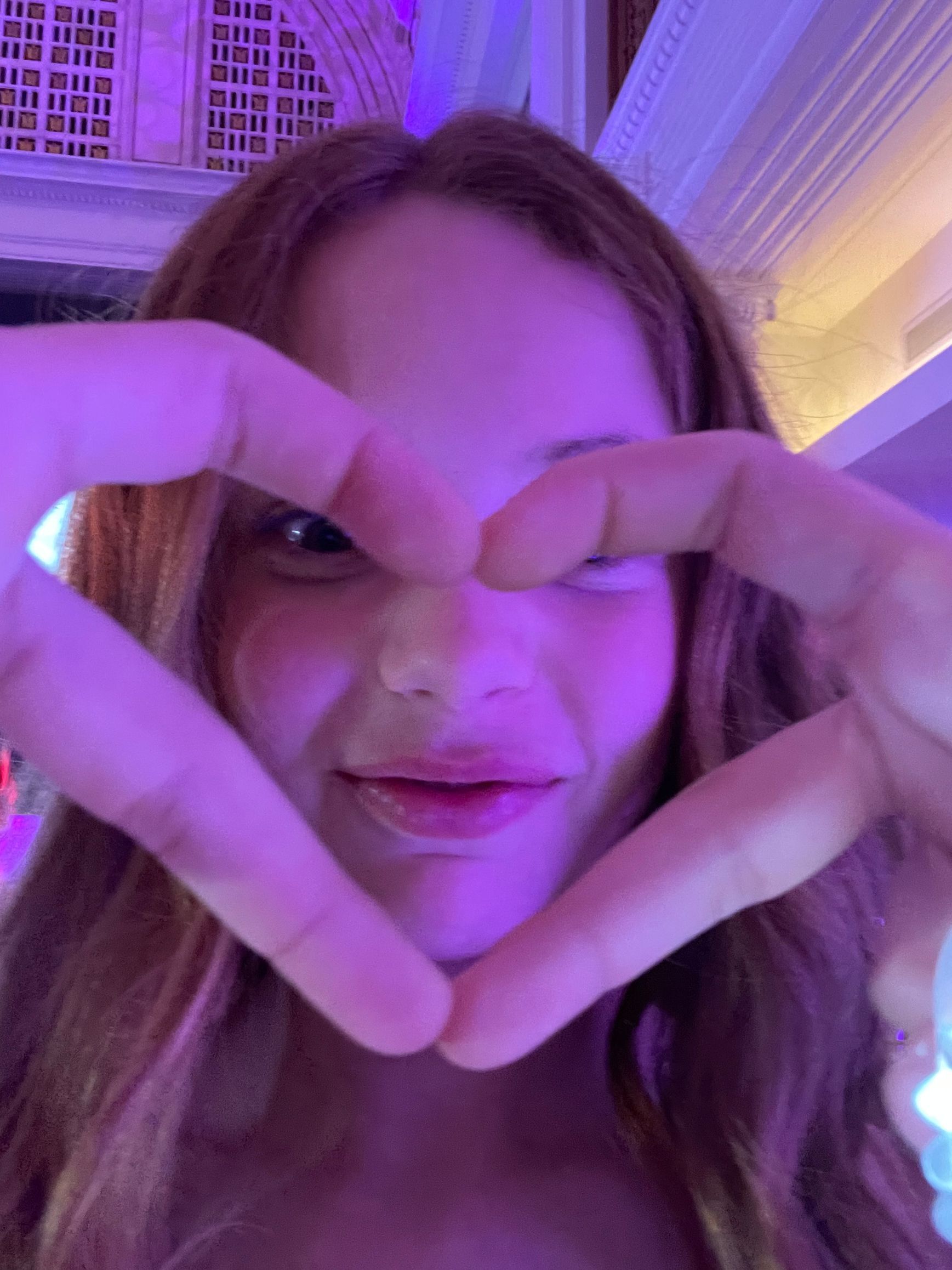 Woman making a heart shape with hands, indoors, lit with purple light, smiling.