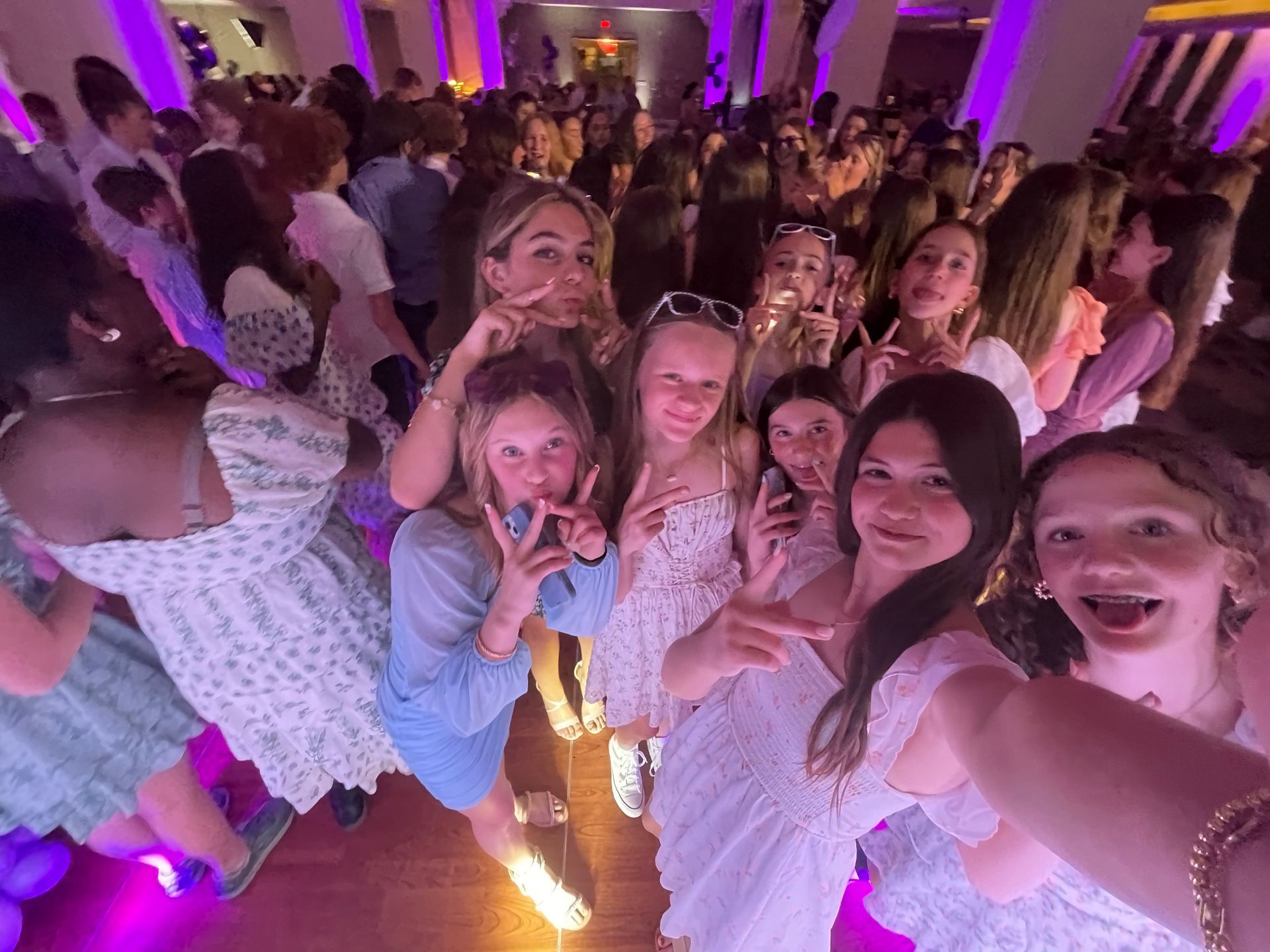 Group of smiling girls take a selfie at a party, with many other people in the background.