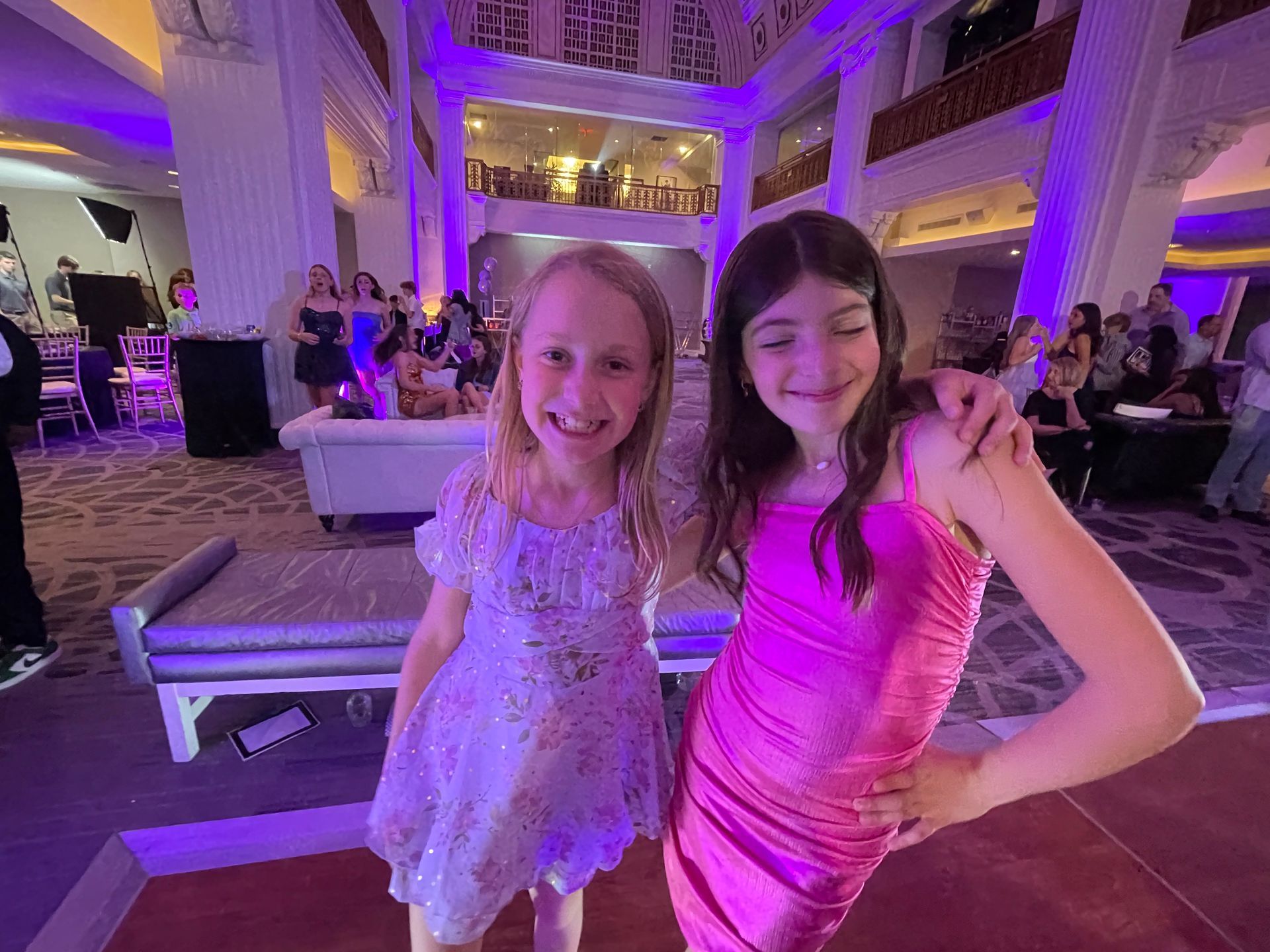 Two girls in dresses smile at the camera in a brightly lit ballroom.
