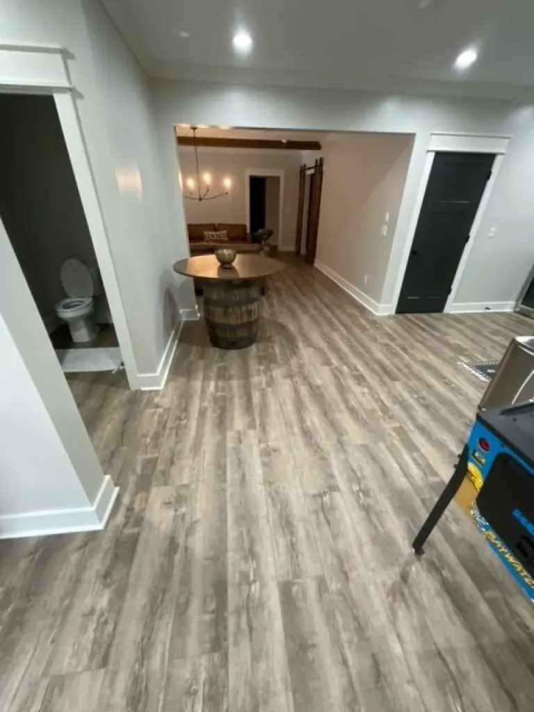 A basement with hardwood floors , a table , a toilet , and a pinball machine.