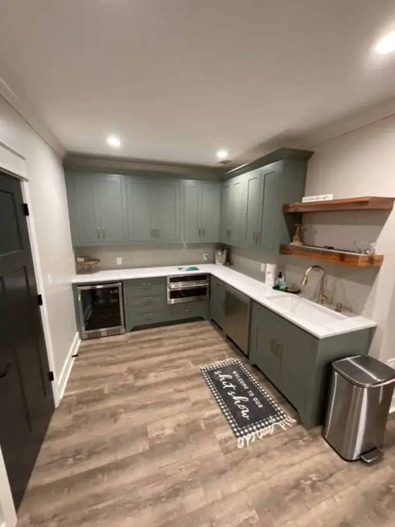 A kitchen with green cabinets , white counter tops , stainless steel appliances and a trash can.