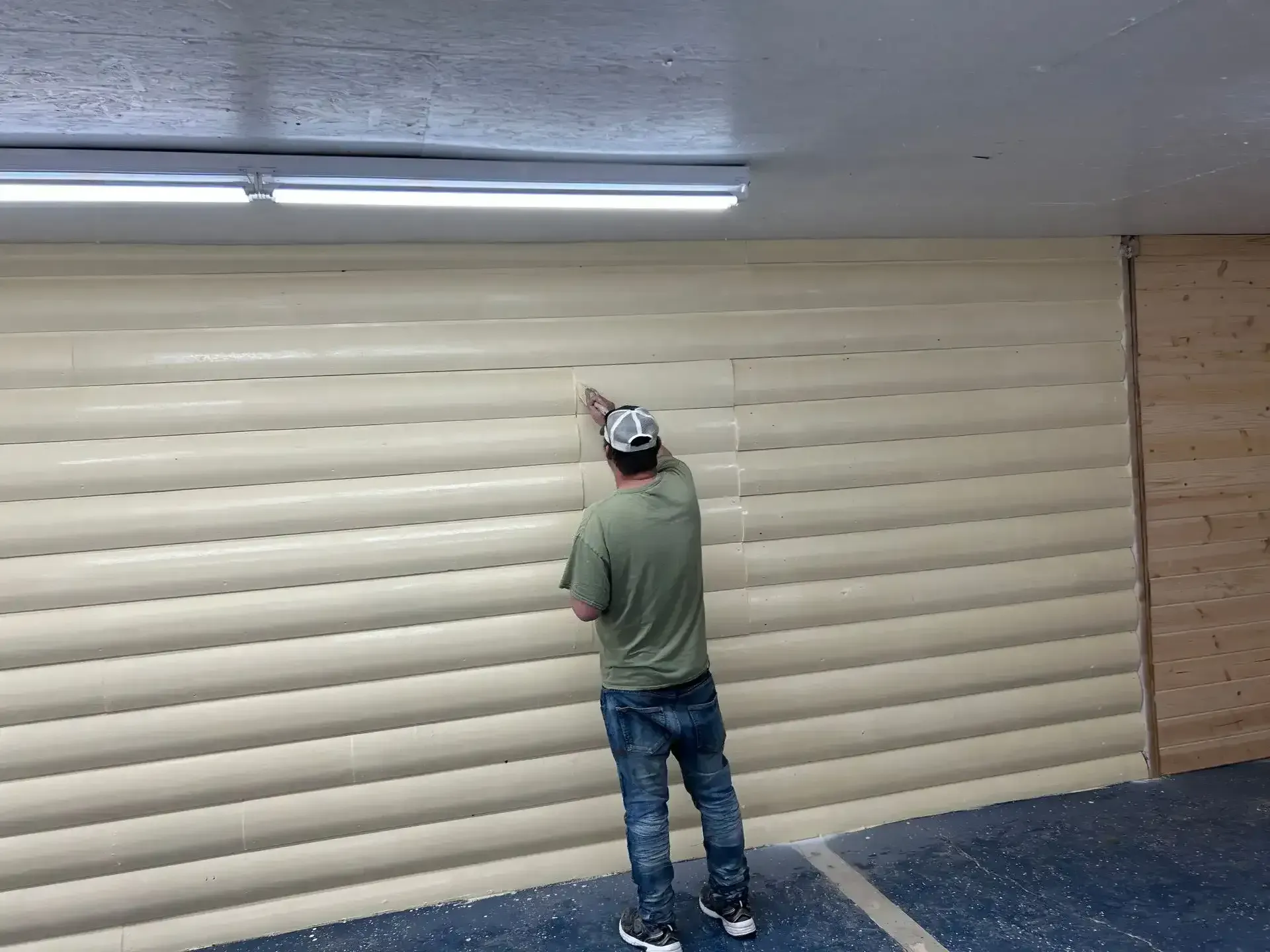 Person in green shirt and jeans working on a beige wall with horizontal ridges, under a fluorescent light.