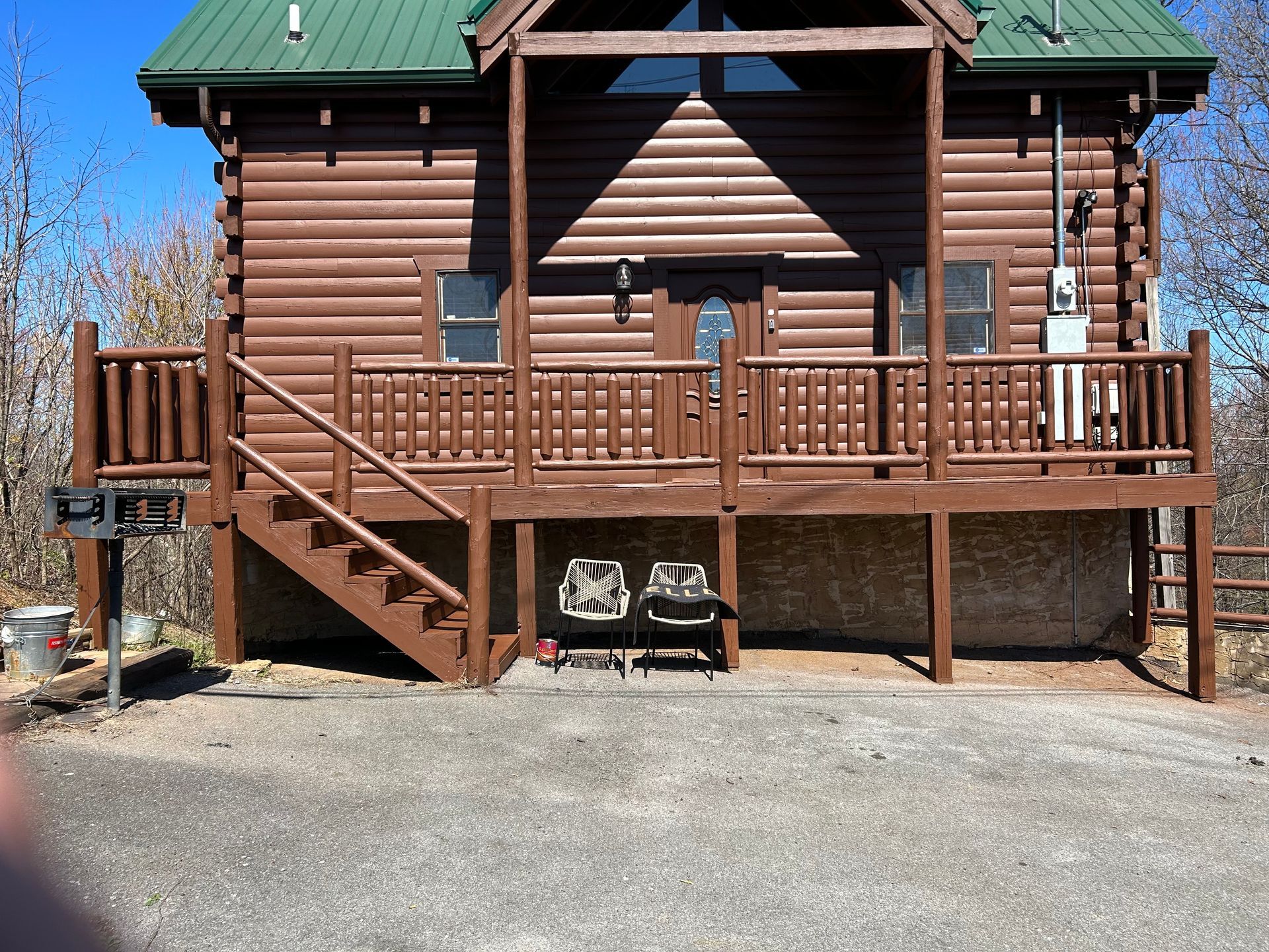 Brown cabin with green roof and wooden deck. Two chairs sit under the deck.