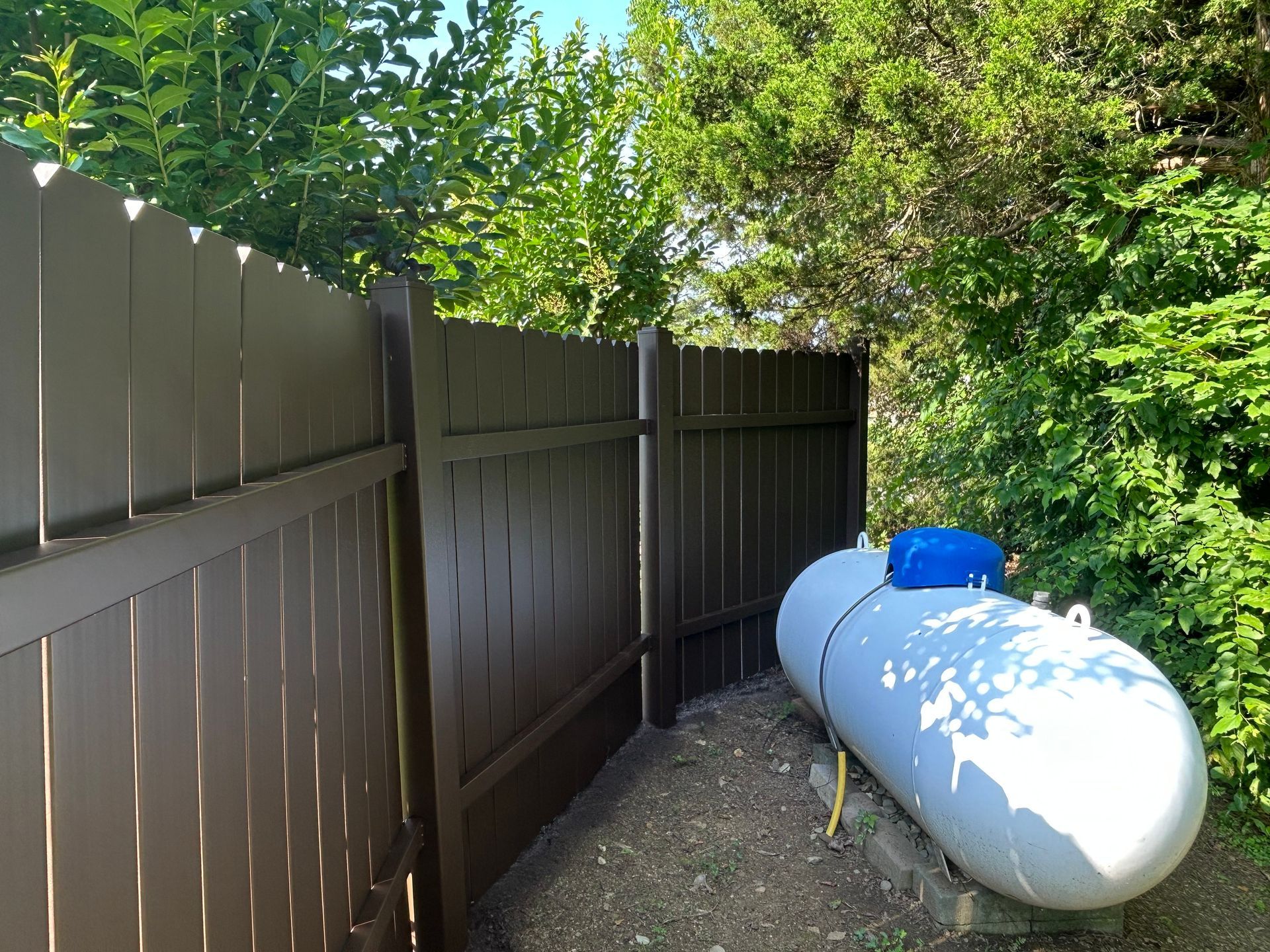 Brown fence encloses a propane tank near trees.