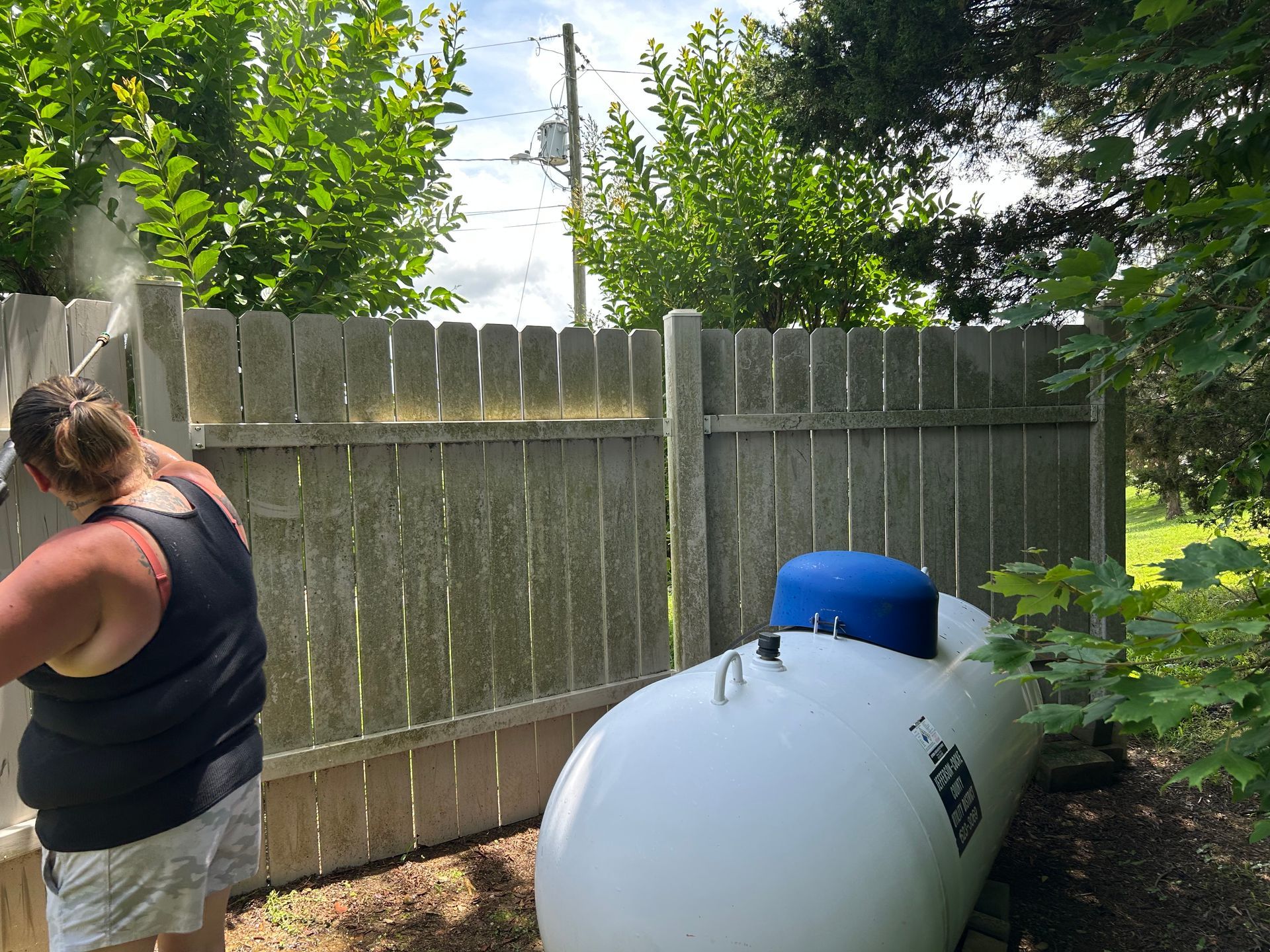 Person pressure washing a weathered wooden fence near a large white propane tank. Green trees and sky in the background.
