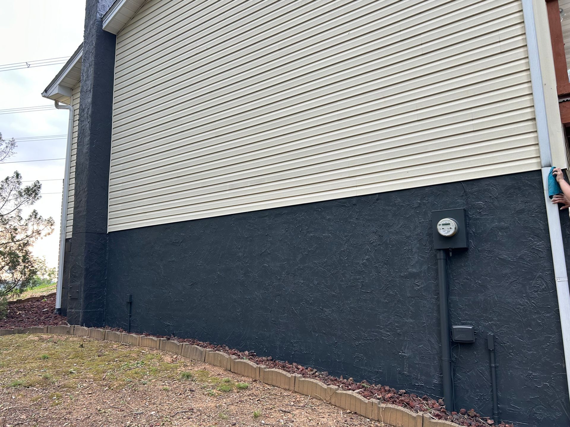 Side of a house with dark gray stucco on the bottom and light tan siding above; electrical box visible.