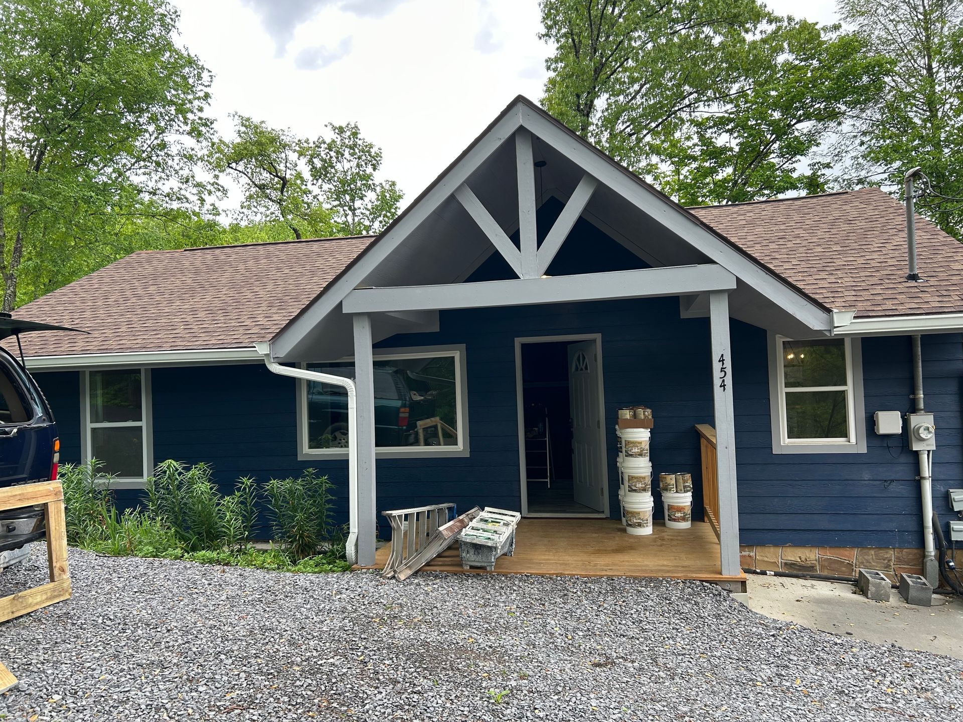 Blue house with white trim, brown roof, and small porch. Gravel driveway and trees in the background.