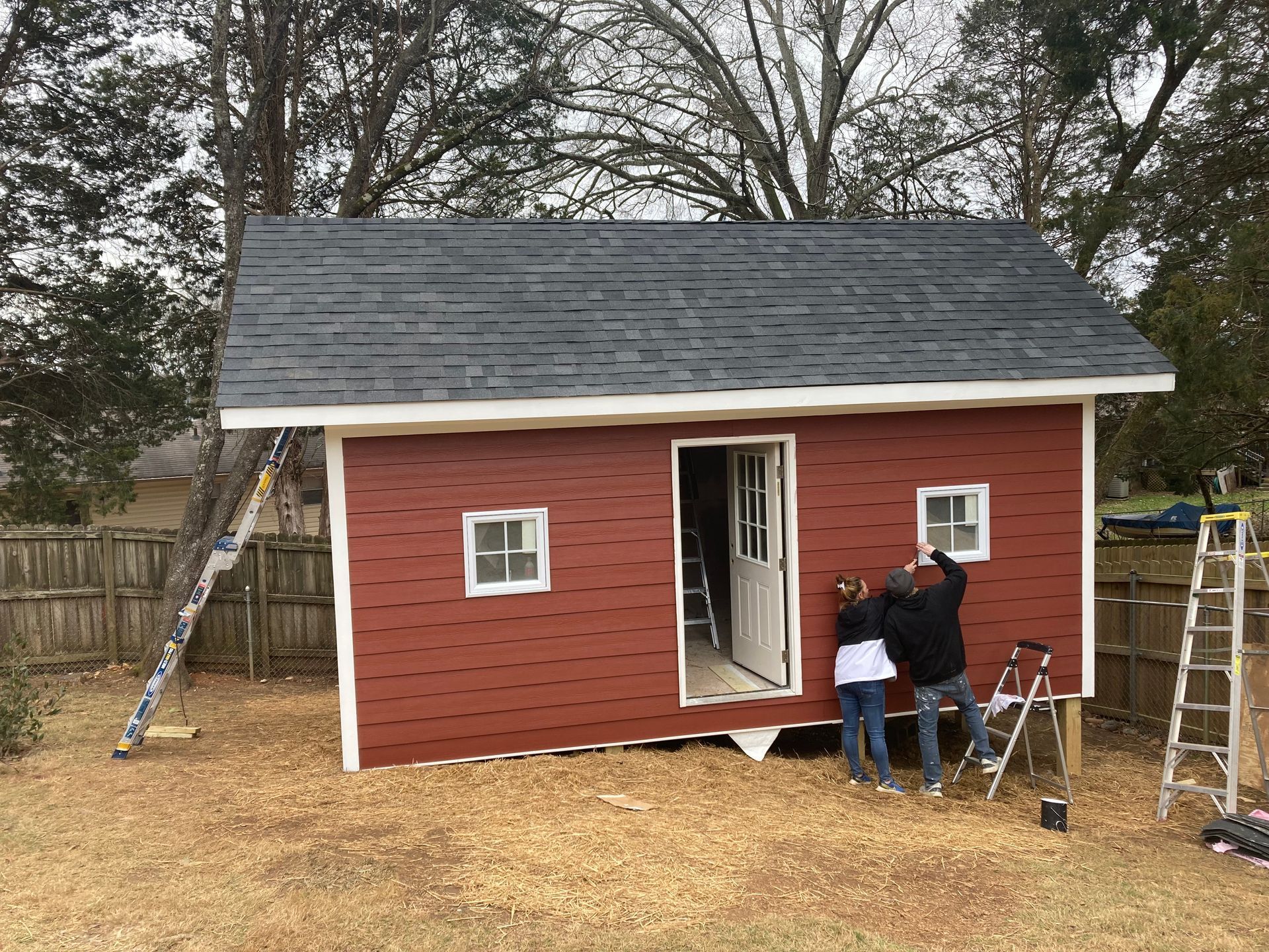 Red shed with gray roof, door open, two people working on it in a yard.
