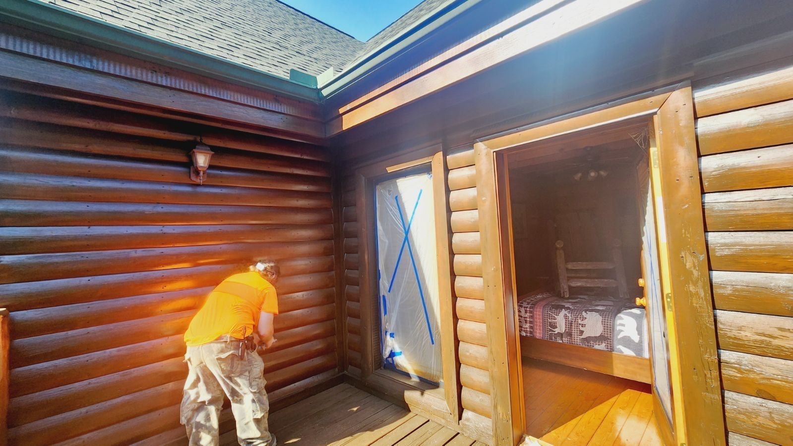 Person painting the exterior of a log cabin; natural wood, door open with paint cans inside.