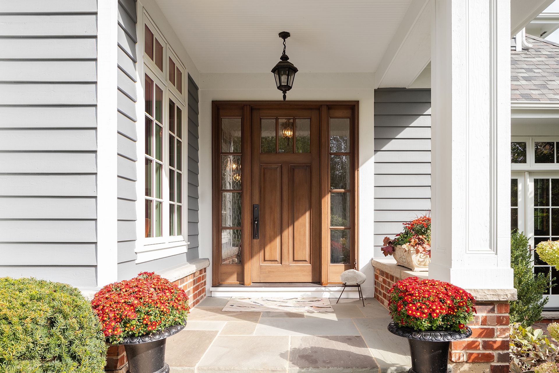 Wooden front door with sidelights, under a covered porch with gray siding and red flowerpots.