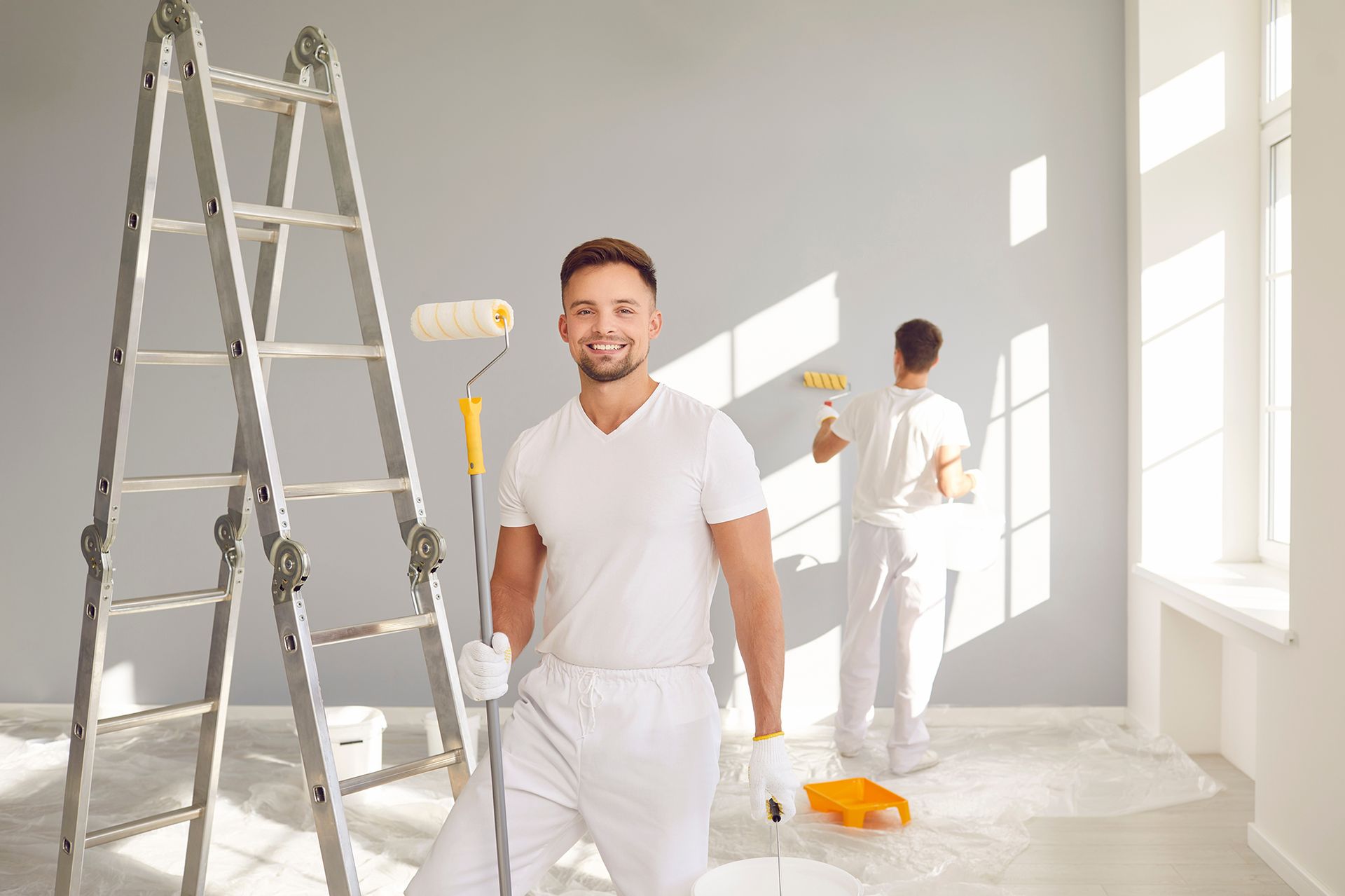 Two painters in white coveralls painting a gray wall; one smiling, holding a roller, the other in the background.