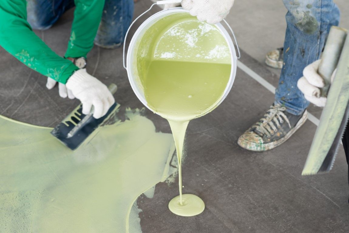 Workers pouring pale green liquid onto a concrete floor during a coating or repair job