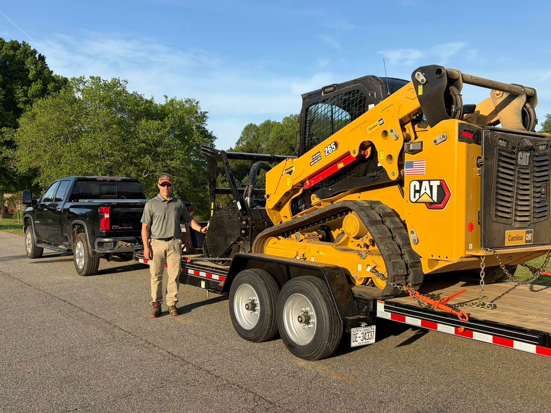 Yellow CAT skid steer on trailer beside black pickup, with a man standing on a road.
