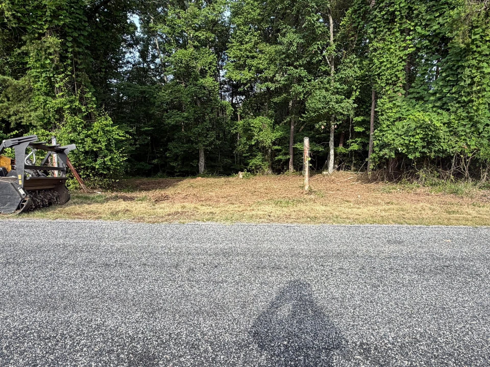 Gravel road beside a wooded lot, with a small excavator partly visible on the left.