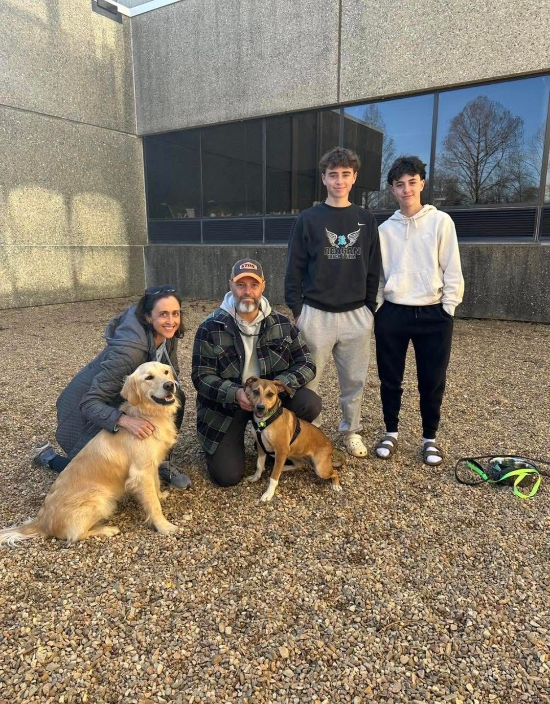 Three people with two dogs posing outside a building on a gravel courtyard