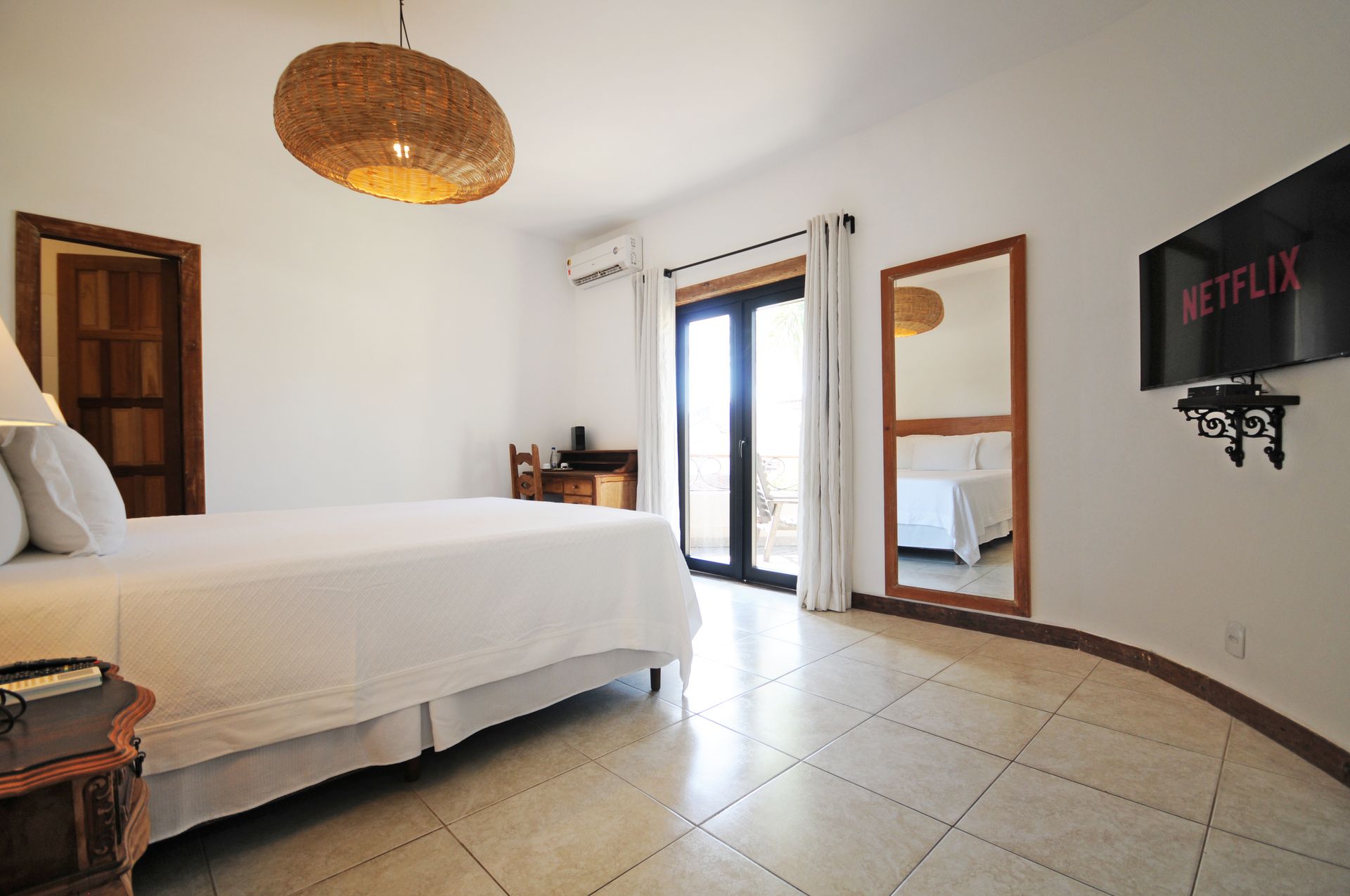 Bedroom with white bedding, wooden door, large mirror, TV, and woven light fixture.