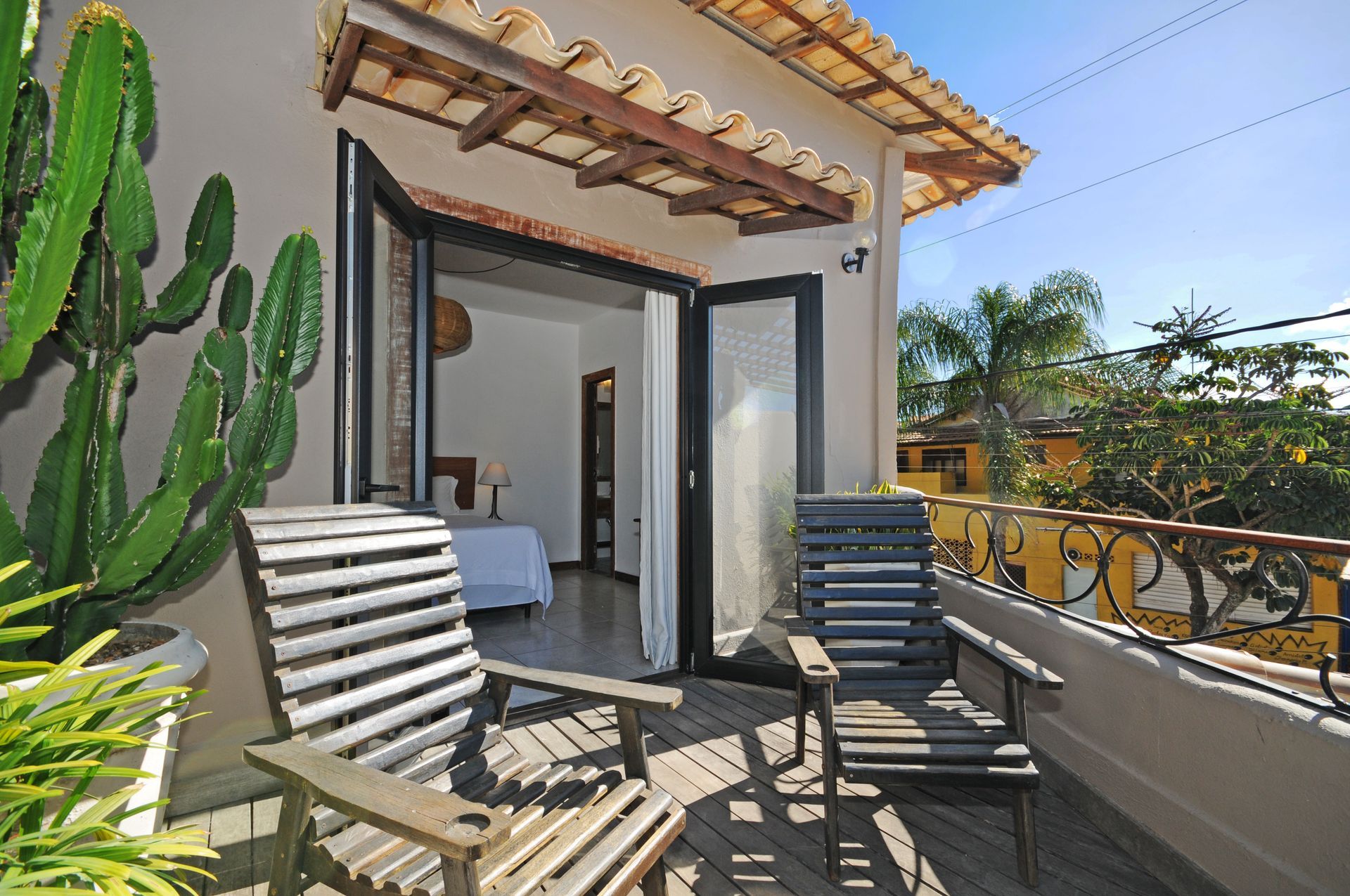 Balcony with wooden chairs, large cactus, open doors to bedroom. Sunny day.