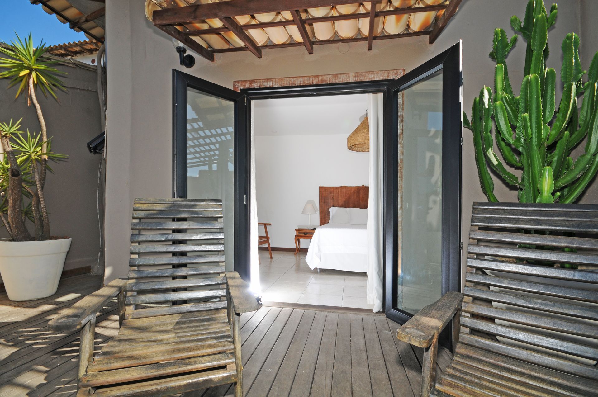 Wooden patio chairs face open doors to a white bedroom. Cactus and potted plant on either side.