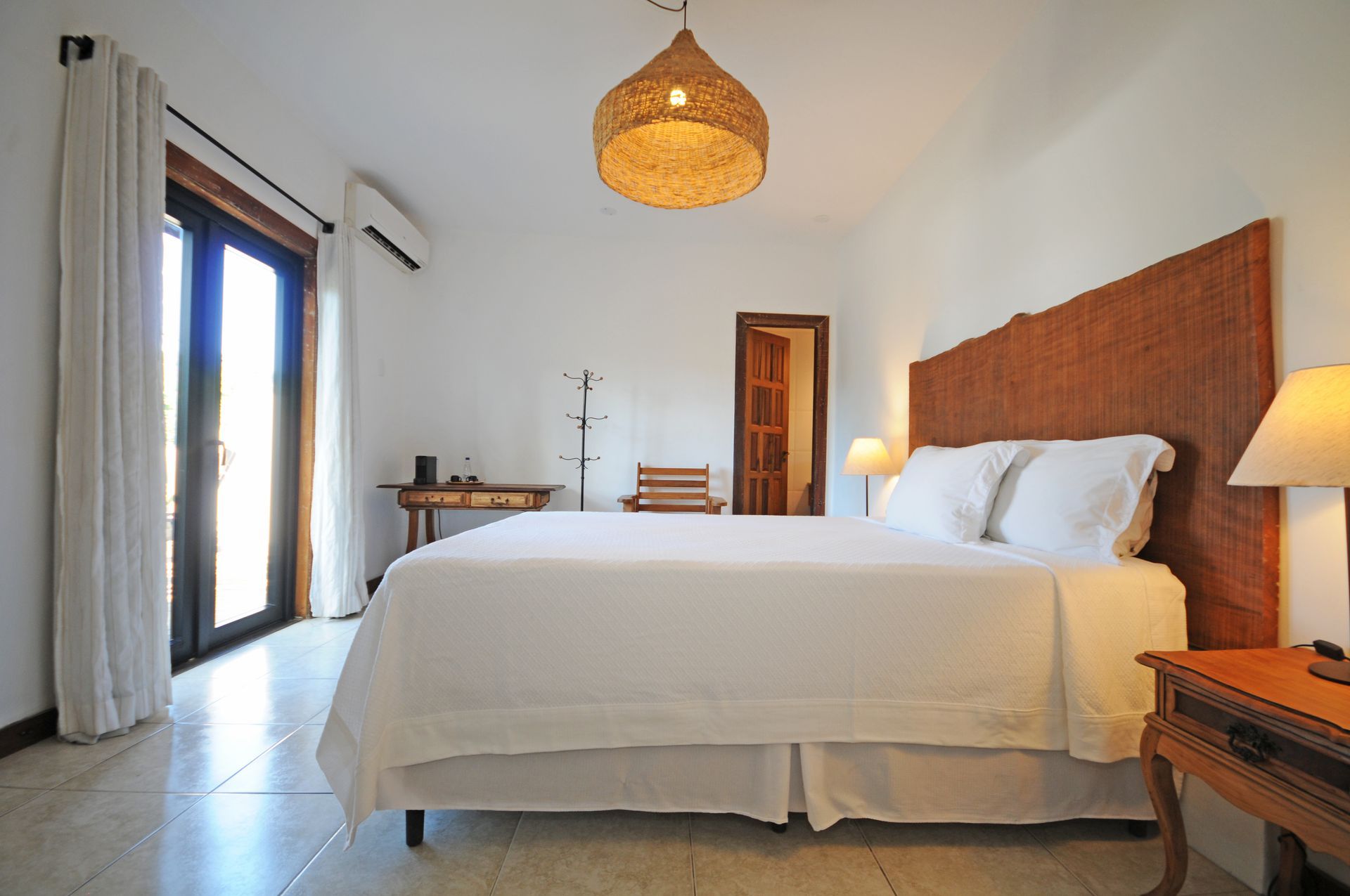 Bedroom with a large bed, woven headboard, and rattan light fixture. White walls, wood accents, and a balcony.