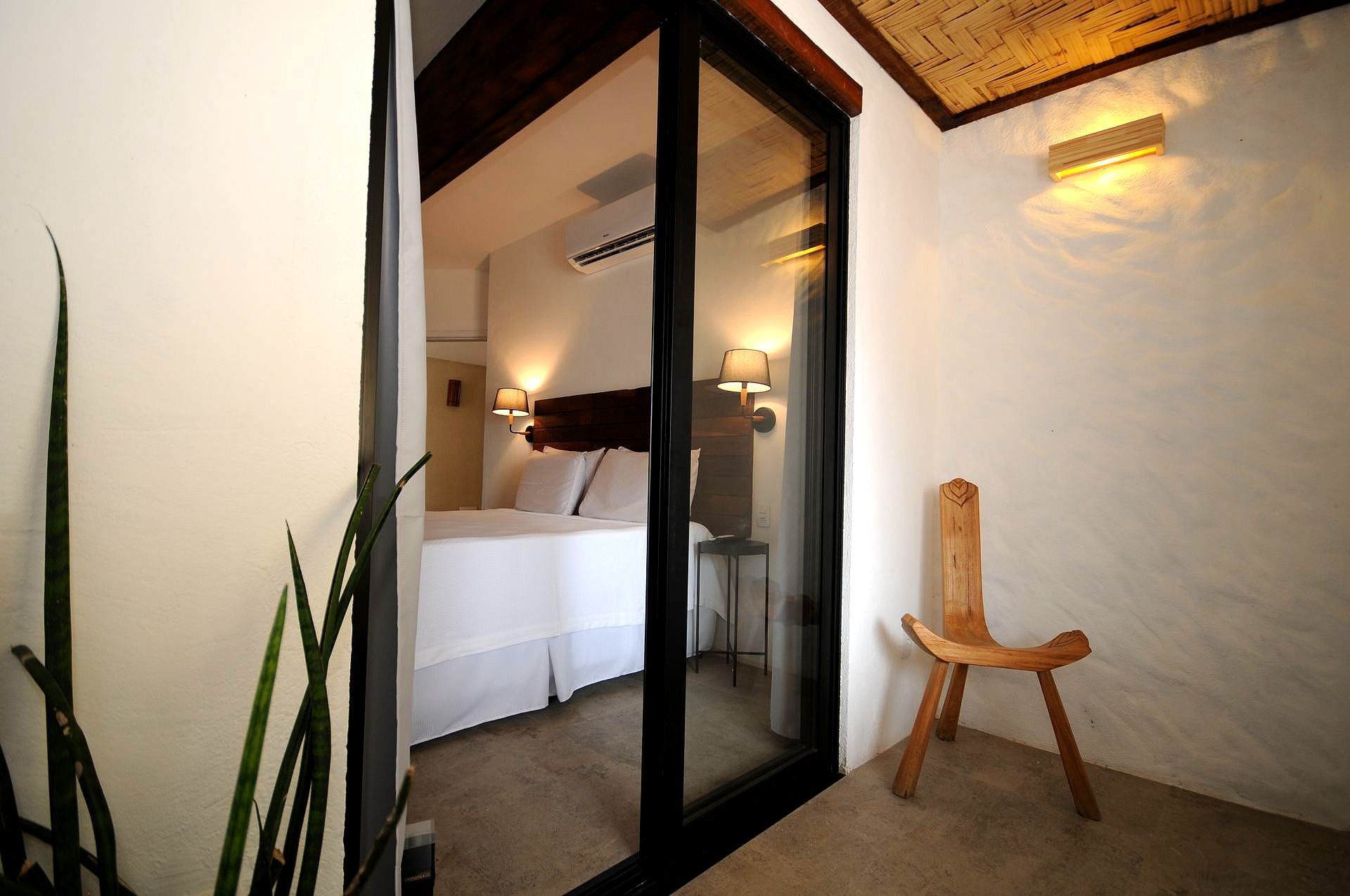 Bedroom interior visible through a glass sliding door. White walls, a wooden chair, and a bed with white linens.