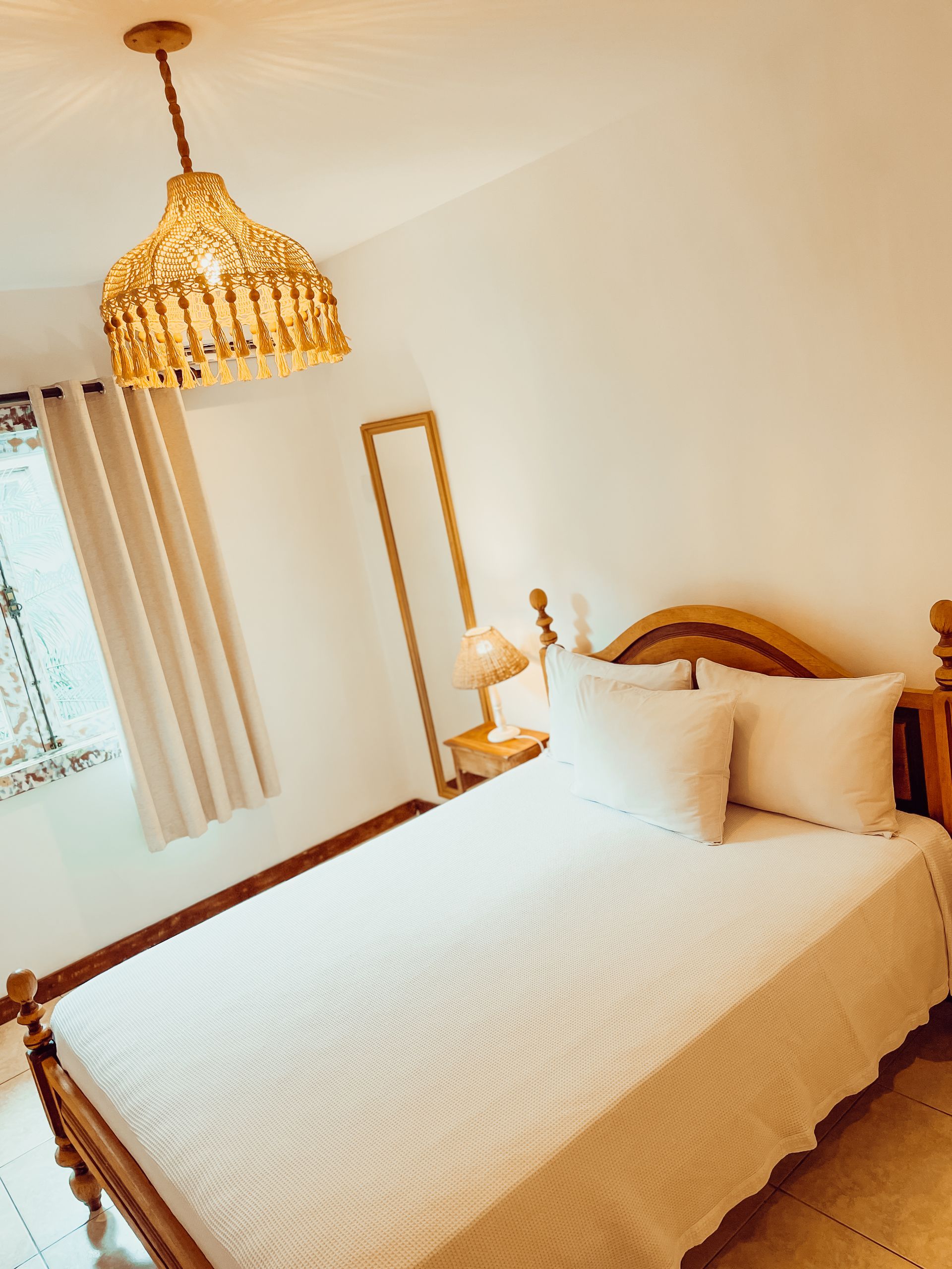 Bedroom with a bed, a lamp, and a tall mirror. White bedding, wooden headboard, and pendant lamp.