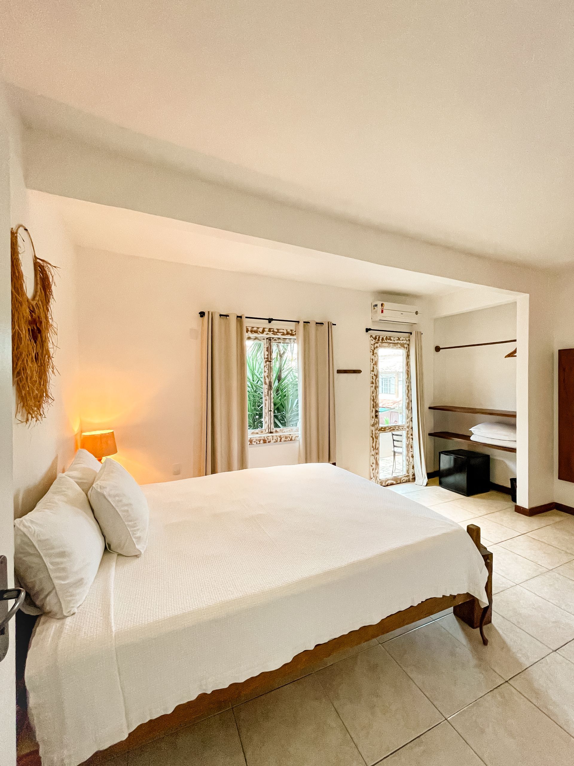 Bright bedroom with a large bed, window, and doorway to a patio. White walls, tile floor, and wooden bed frame.