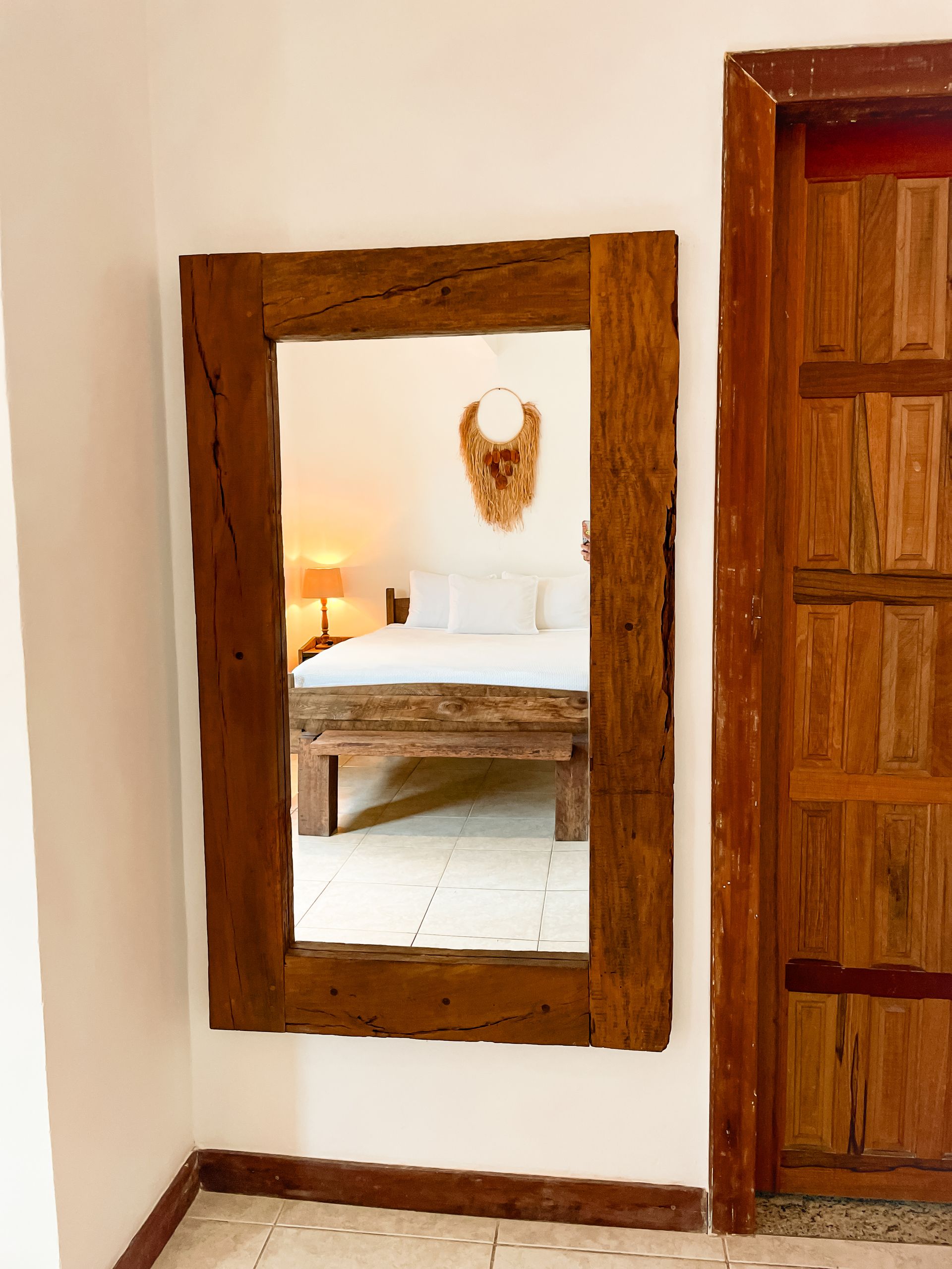 Large, rectangular mirror with a thick, wooden frame reflects a bedroom with a bed and a decorative wall hanging.
