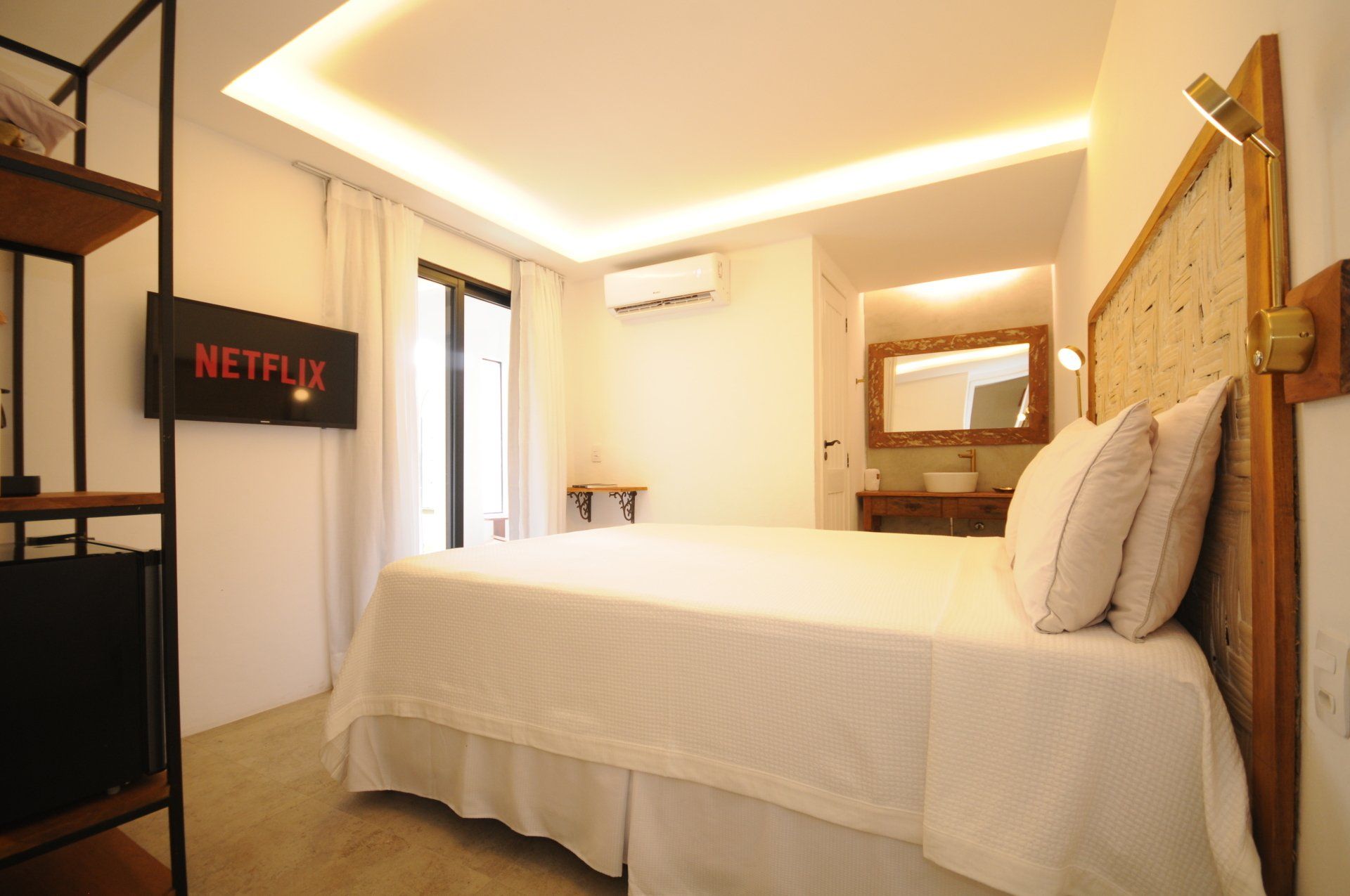 Bedroom with bed, TV, shelves, and a doorway to a bathroom. Warm lighting and white walls.
