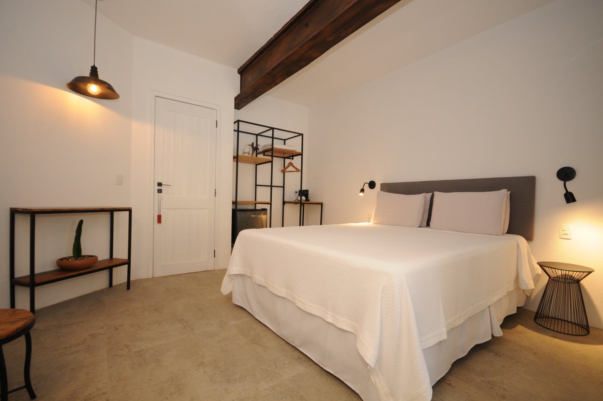 Bedroom with a bed, a shelf, and hanging lights; white walls, wooden beams, concrete floor.