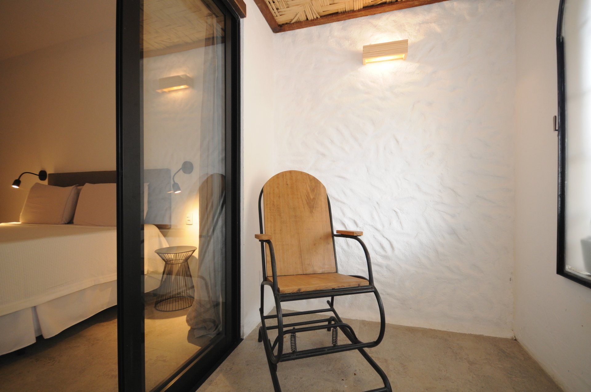 Small balcony with wooden rocking chair, next to a bedroom with bed. White textured walls.
