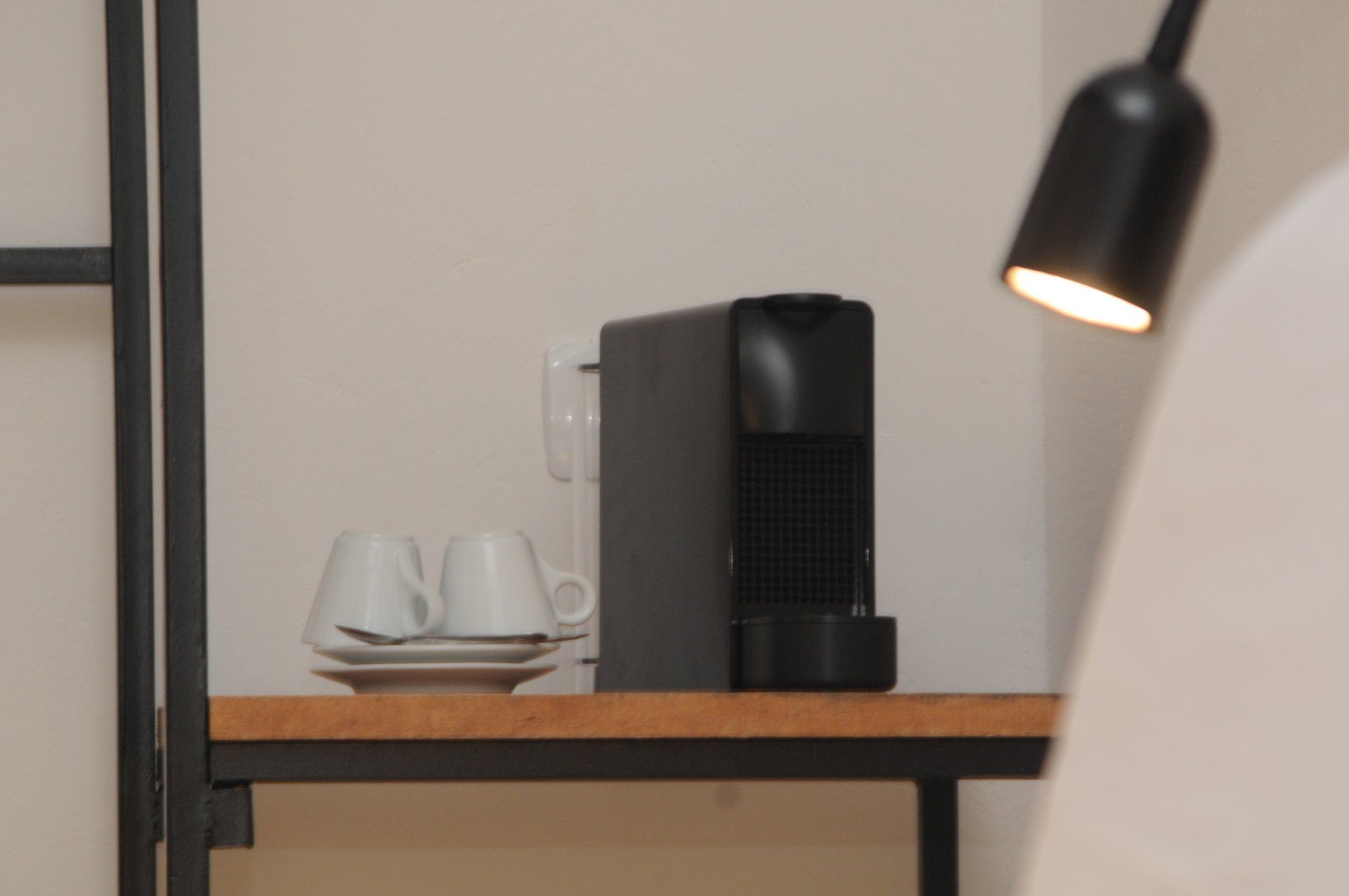 Coffee machine, cups, saucer on a wooden shelf, black lamp overhead. White wall background.