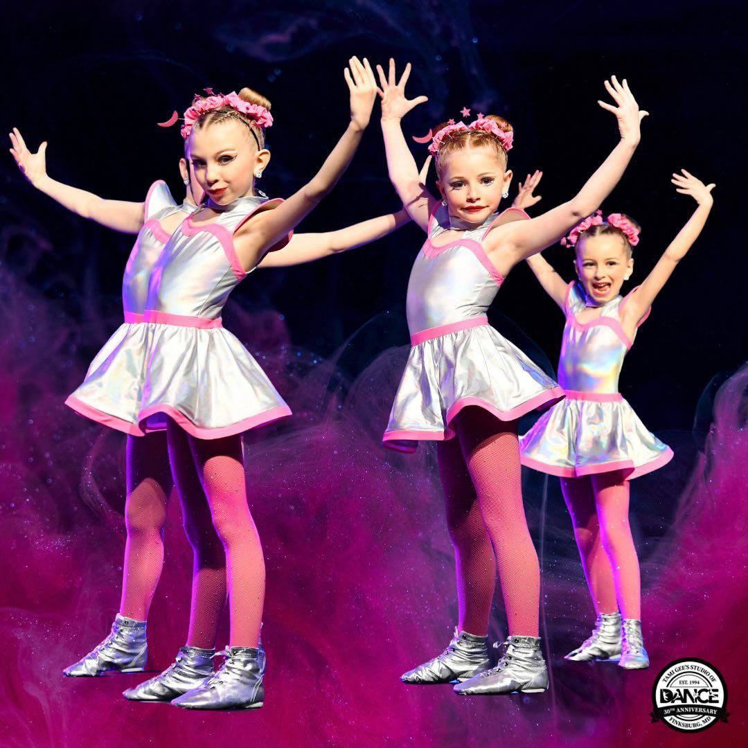 Three little girls are dancing on a stage with their arms in the air.