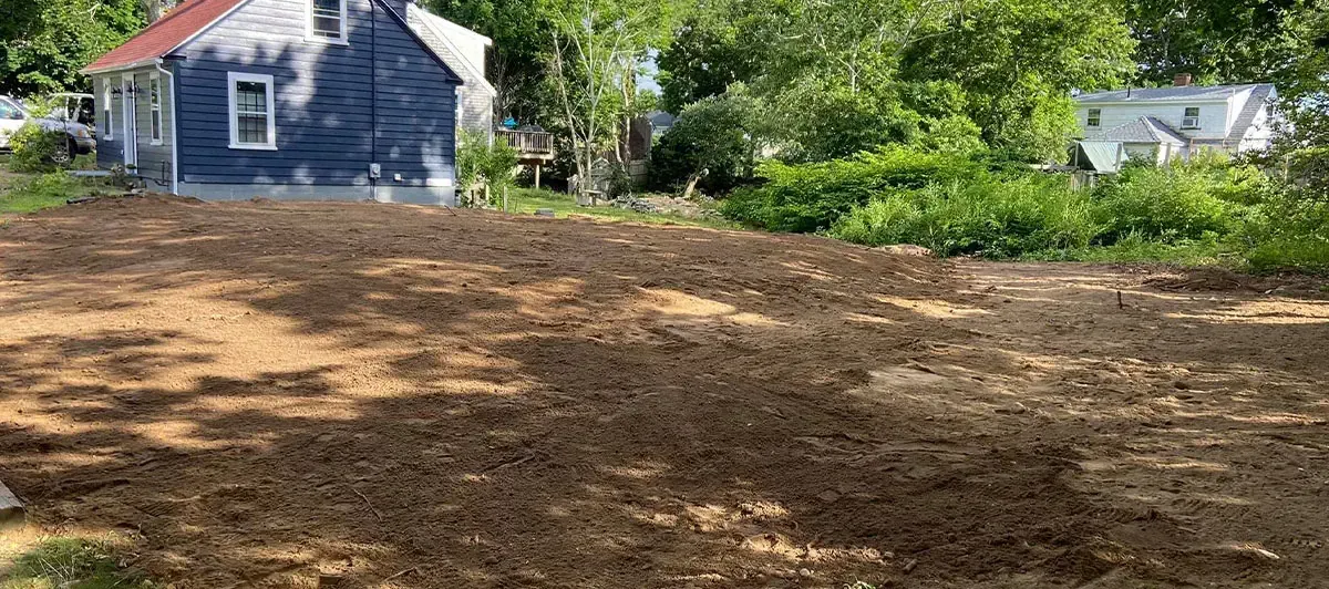 A leveled dirt yard in front of a blue house with red roof. Trees and another house are in the background.