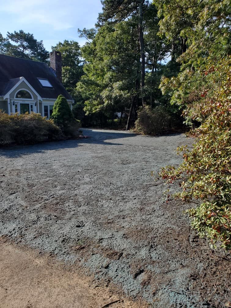 Bare, gravel-covered area in front of a house, surrounded by trees and bushes under a sunny sky.