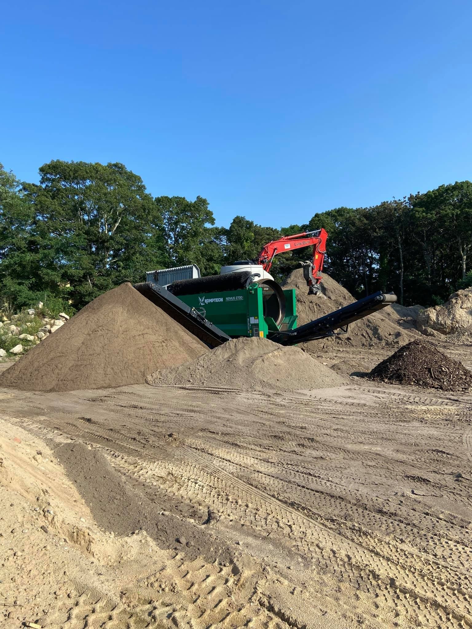 Construction site with soil piles, a green screening machine, and an excavator under a blue sky.