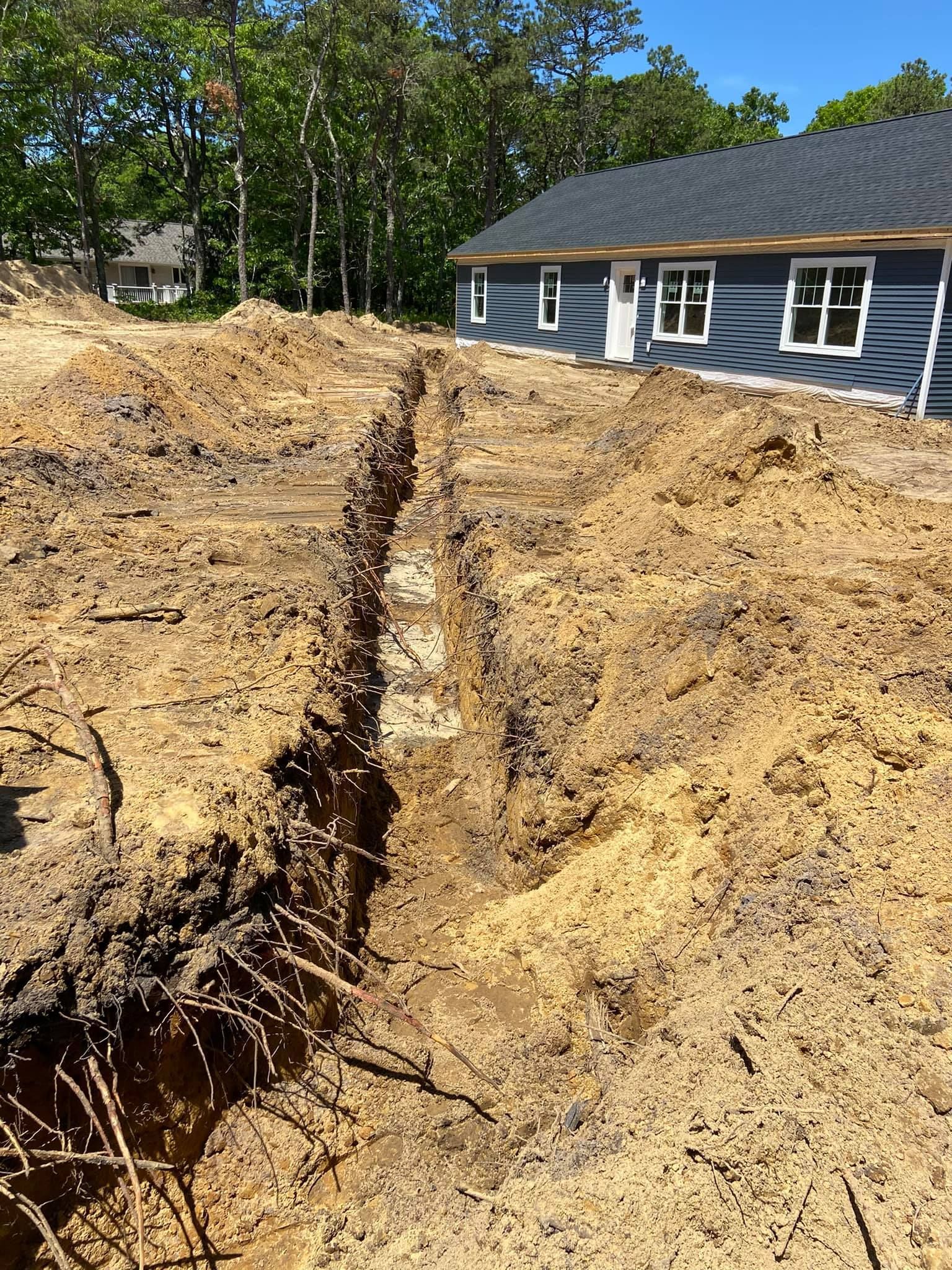 Trench dug in dirt in front of a blue-sided house with a dark roof and white windows, under a blue sky.