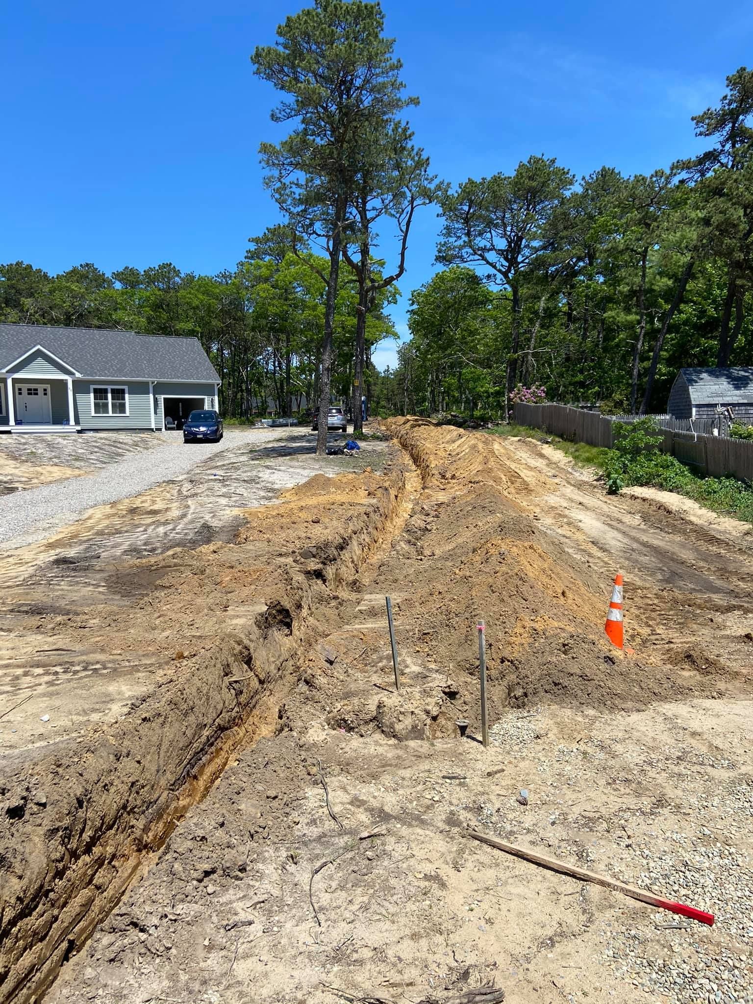 Dirt construction site with trenches, stakes, and orange cone. A completed house and trees in background.