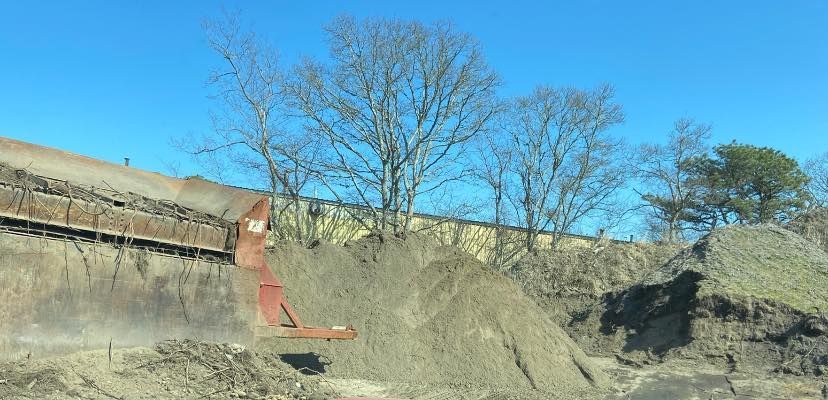 An industrial site with a conveyor belt depositing soil onto a pile, trees, blue sky.