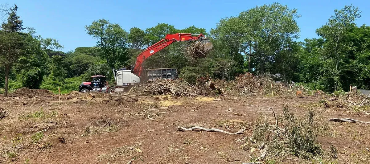 An excavator and tractor clearing land of trees and debris under a clear blue sky.