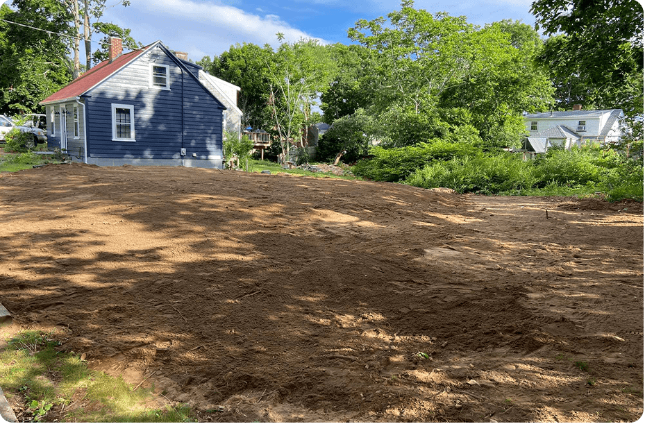 Cleared lot with brown soil in front of blue house with red roof, trees, and other houses.