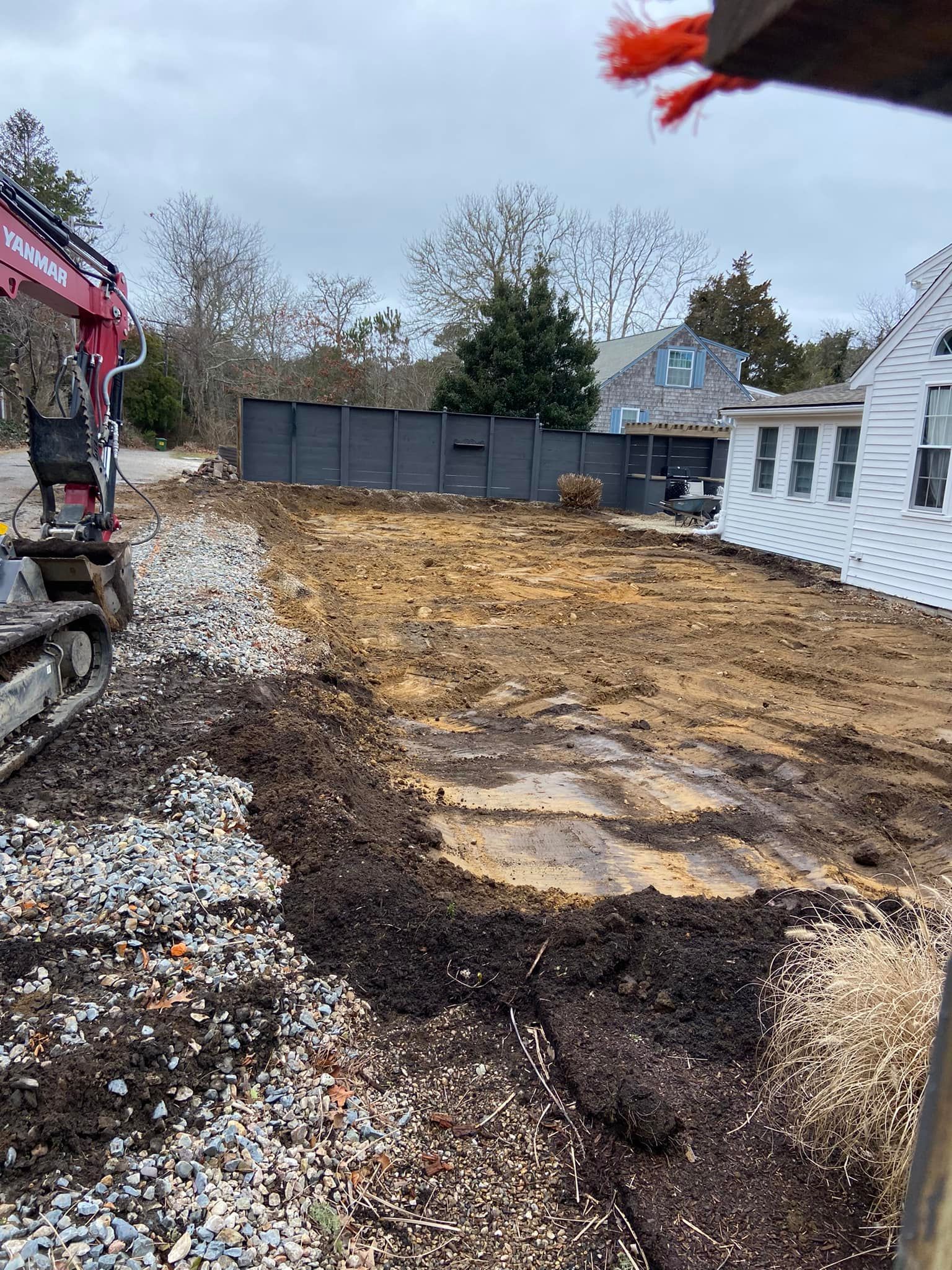 Excavated construction site with an excavator, soil, gravel, and nearby houses. Cloudy day.