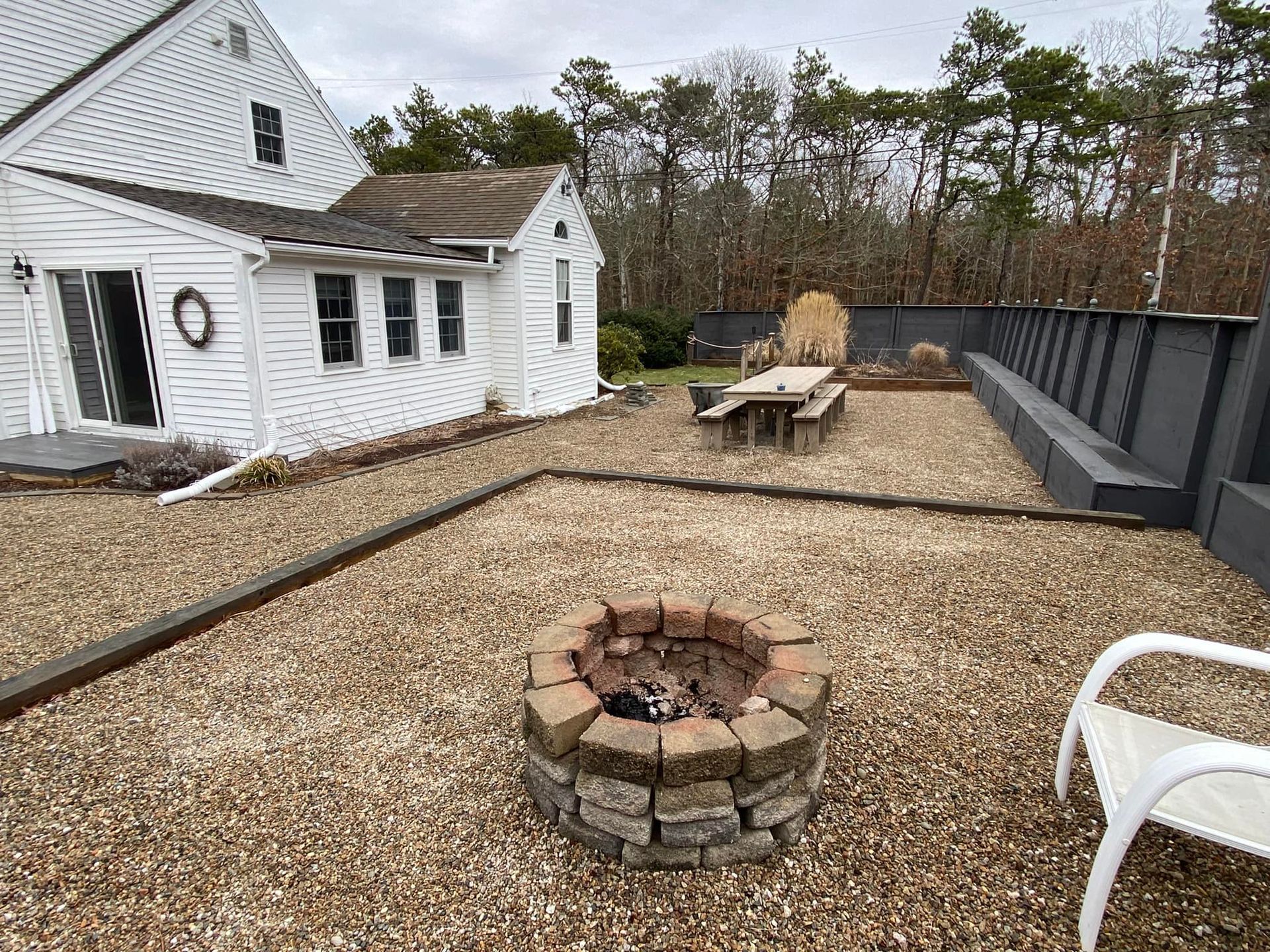 Backyard patio with fire pit, gravel ground, picnic table, and white house. Overcast sky.
