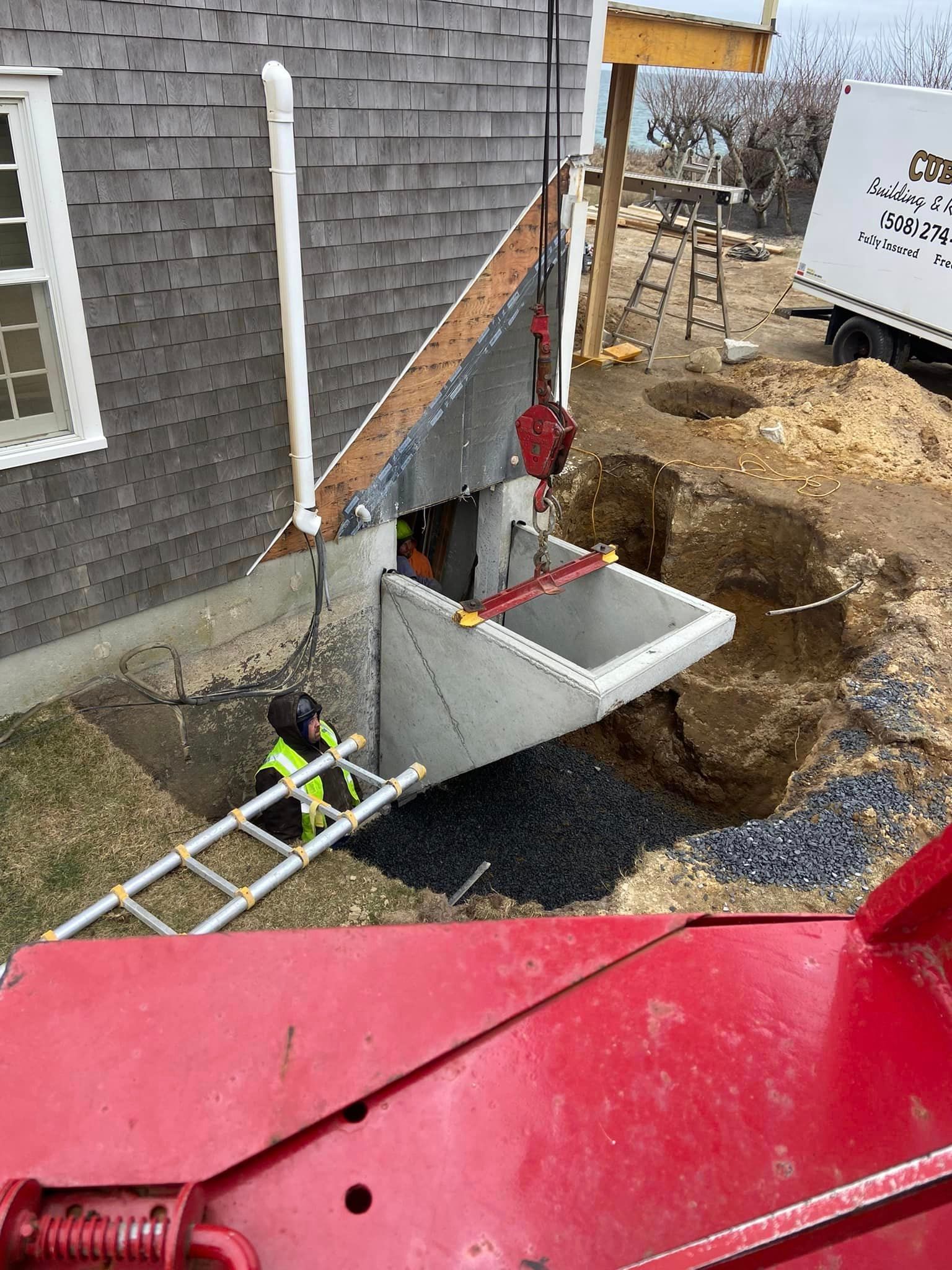 Construction site: Crane lifting a concrete structure into a foundation opening, with worker in safety vest nearby.