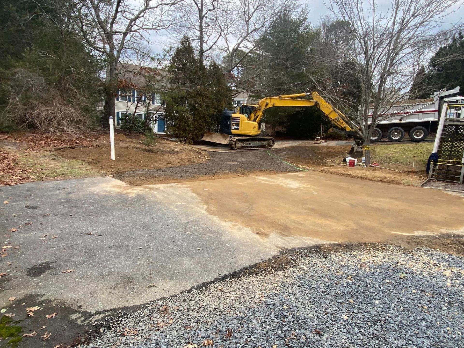 Excavator on a construction site; dirt and gravel driveway. Semi-truck and house in the background.
