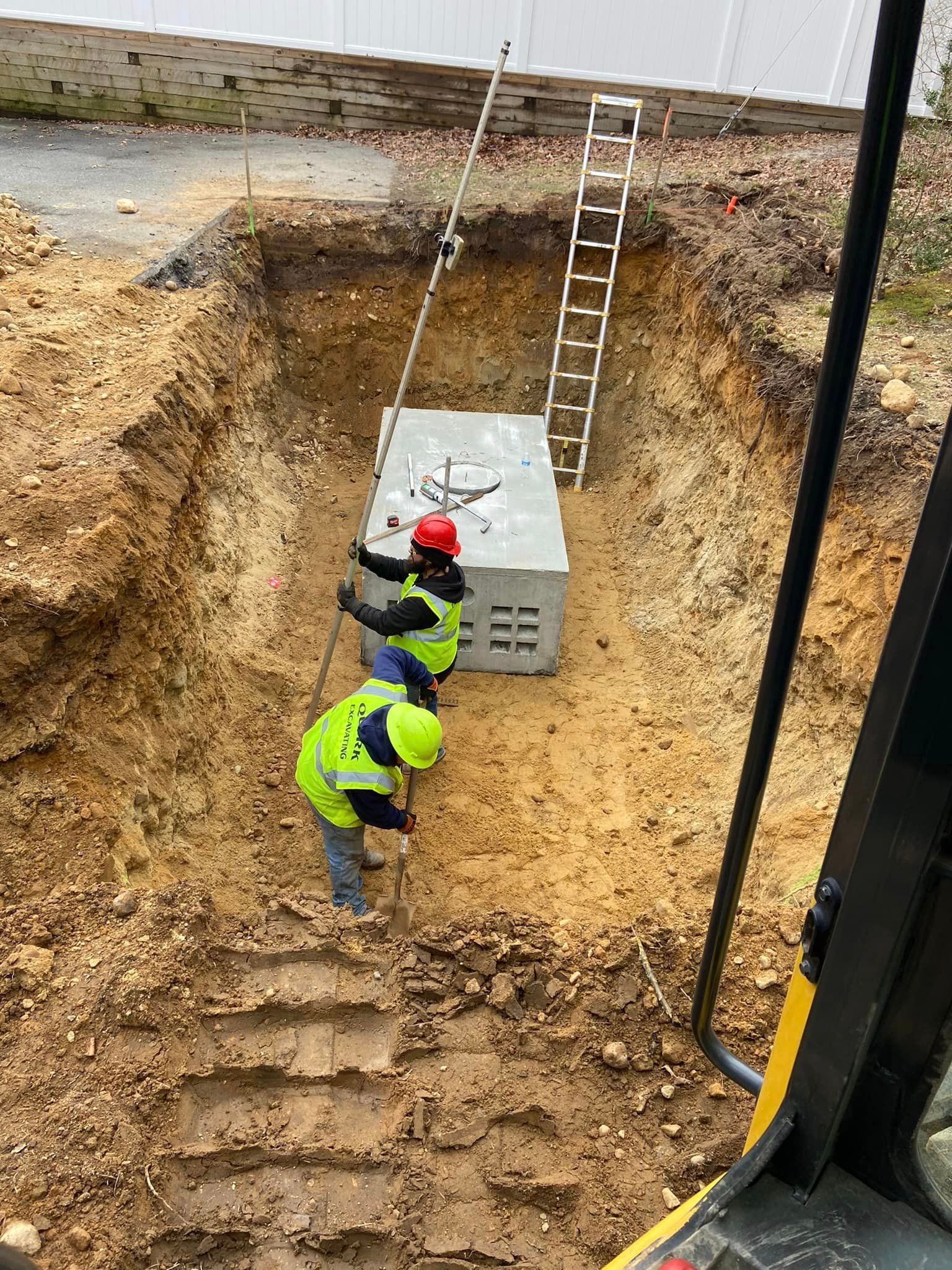 Two workers in safety vests and hard hats lowering a large metal box into a deep pit. A ladder leans nearby.