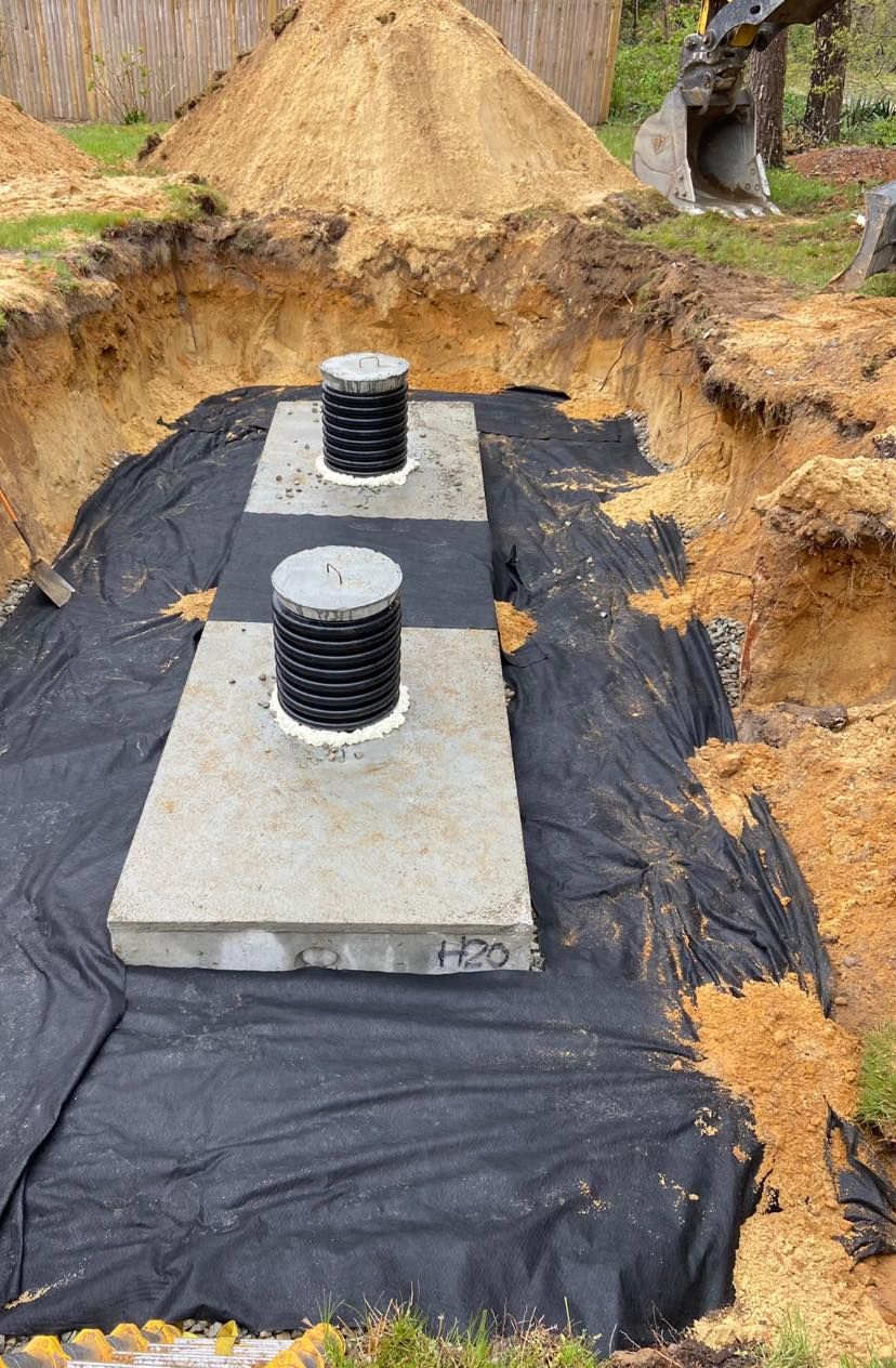 A rectangular concrete structure with two black capped openings in a dug-out hole, lined with black fabric.