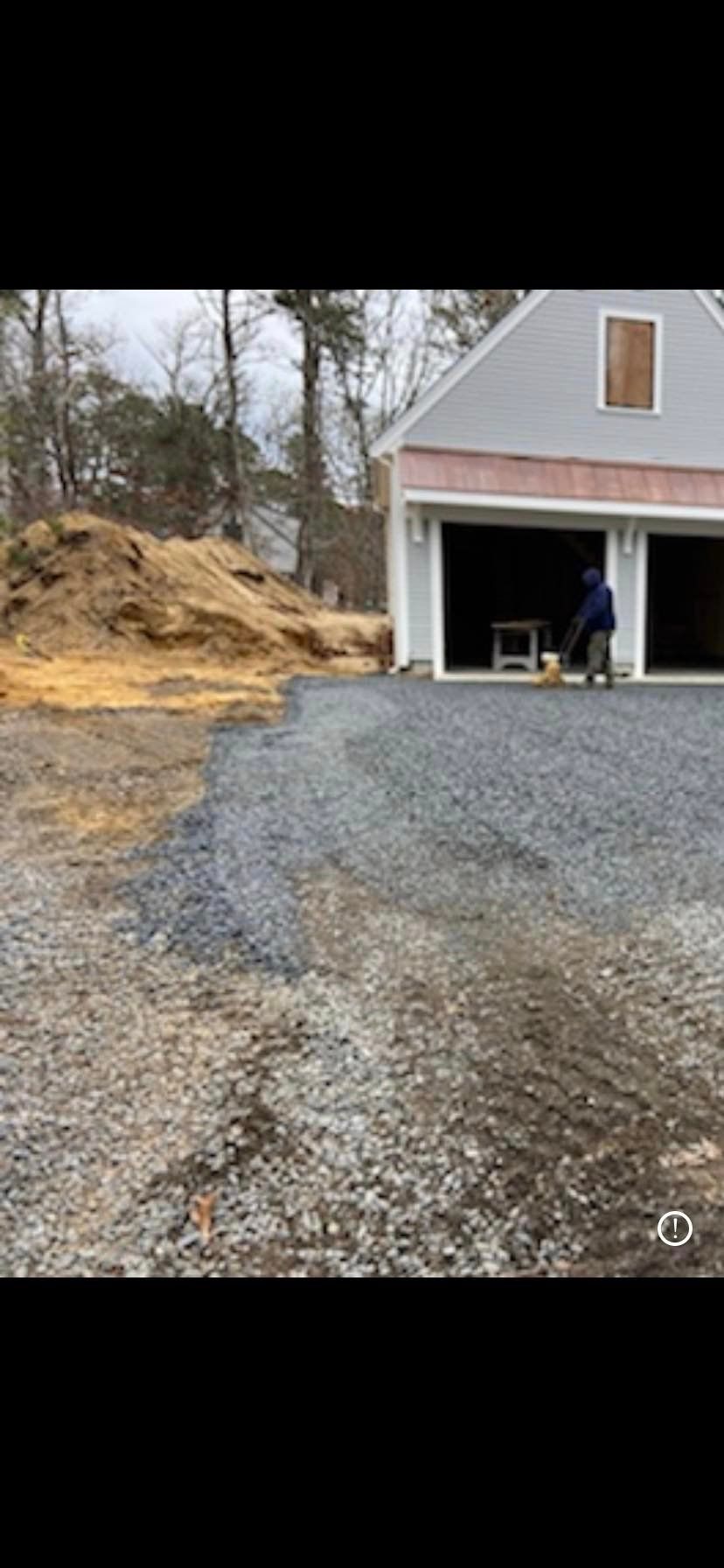 A gravel driveway leads to a two-car garage. A person works in the open garage. A large pile of dirt is nearby.