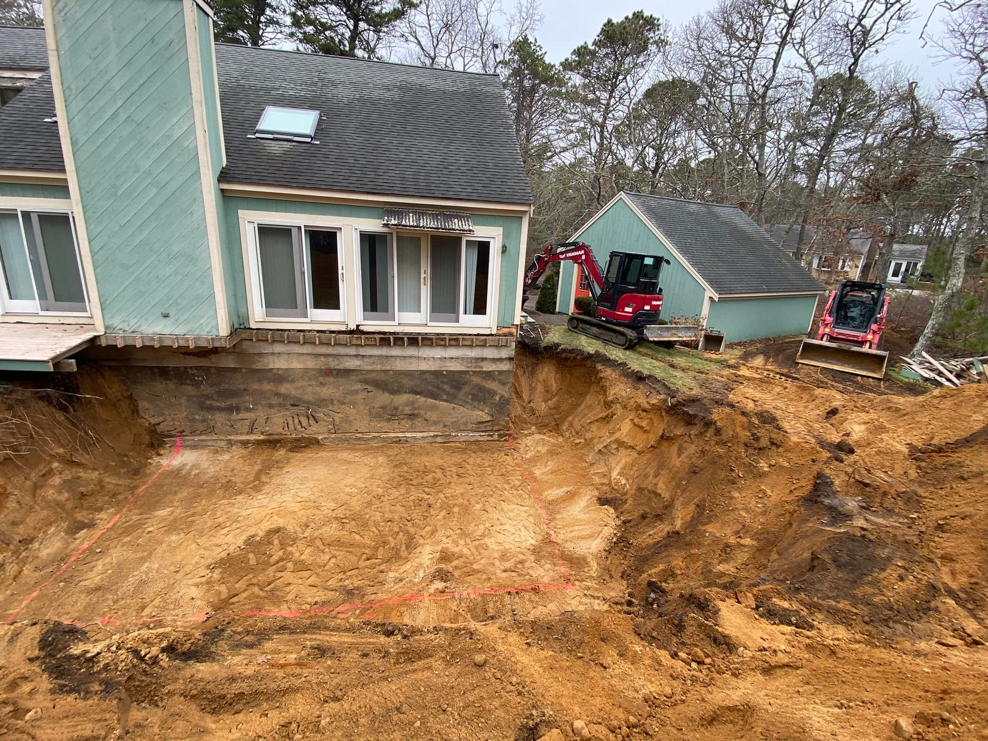 Construction site with house, shed, and excavation. Two red excavators work near a building.