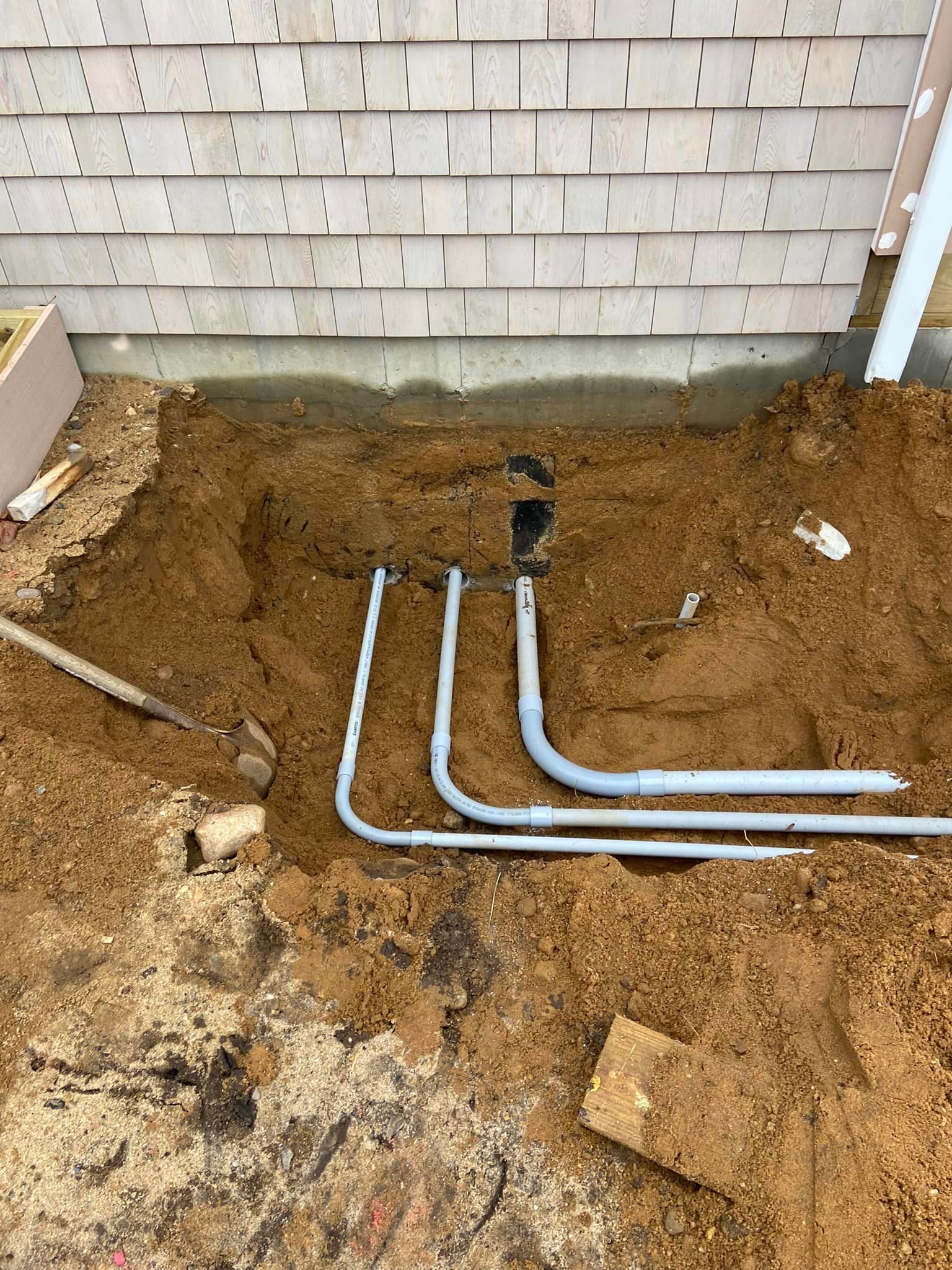 Gray electrical conduits emerging from a trench next to a wooden building.