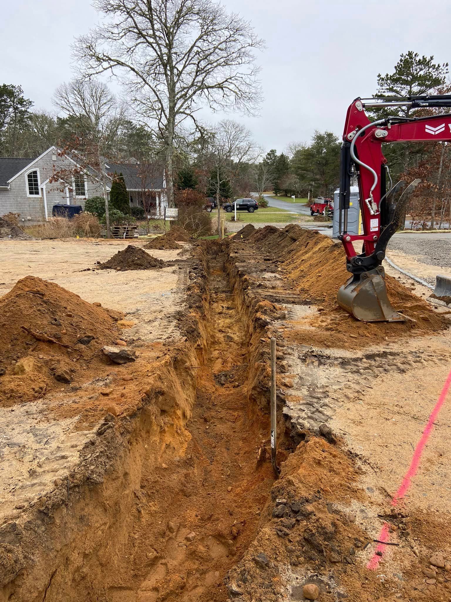 Trench dug in dirt, red excavator, residential area, overcast sky.